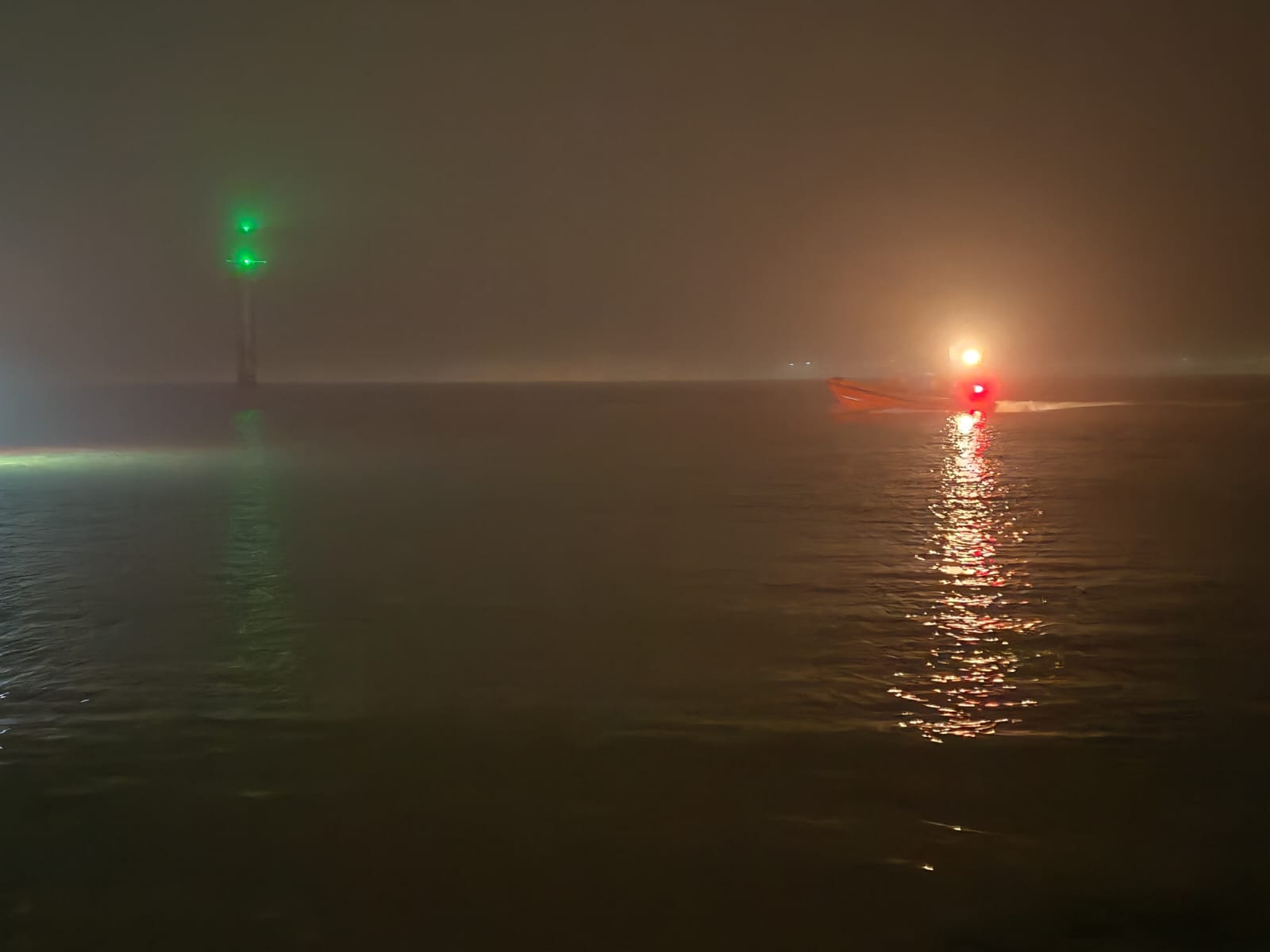 A slightly unclear photo of the river Mersey from the shore. A groyne marker can be seen to the left of the image, with illuminated green top. The lifeboat is off to the right, lit by its navigation lights. A very foggy night.