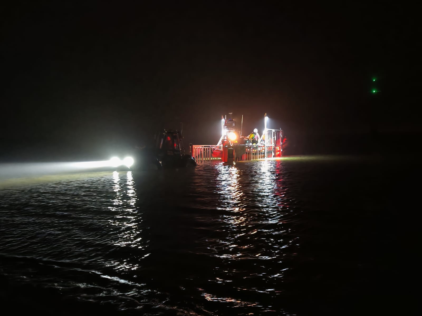 The lifeboat has recovered into the carriage in the dark. The river, sky and tractor can barely be seen, with light only coming from the tractor headlights and the carriage mast lights, illuminating the deck of the lifeboat.
