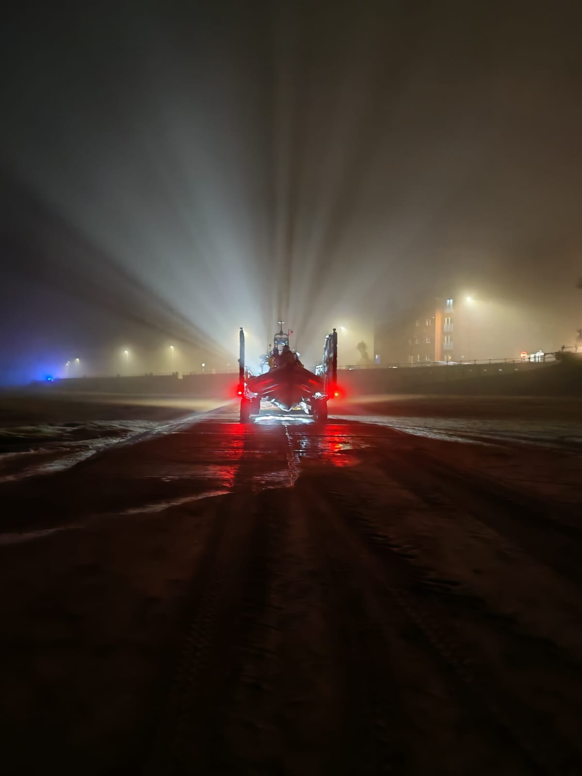 New Brighton RNLI's tractor, carrying the lifeboat, is heading toward the photographer, launching on service. In the fog, beams of light can be seen shining out from the lights behind into the sky. Colour image.