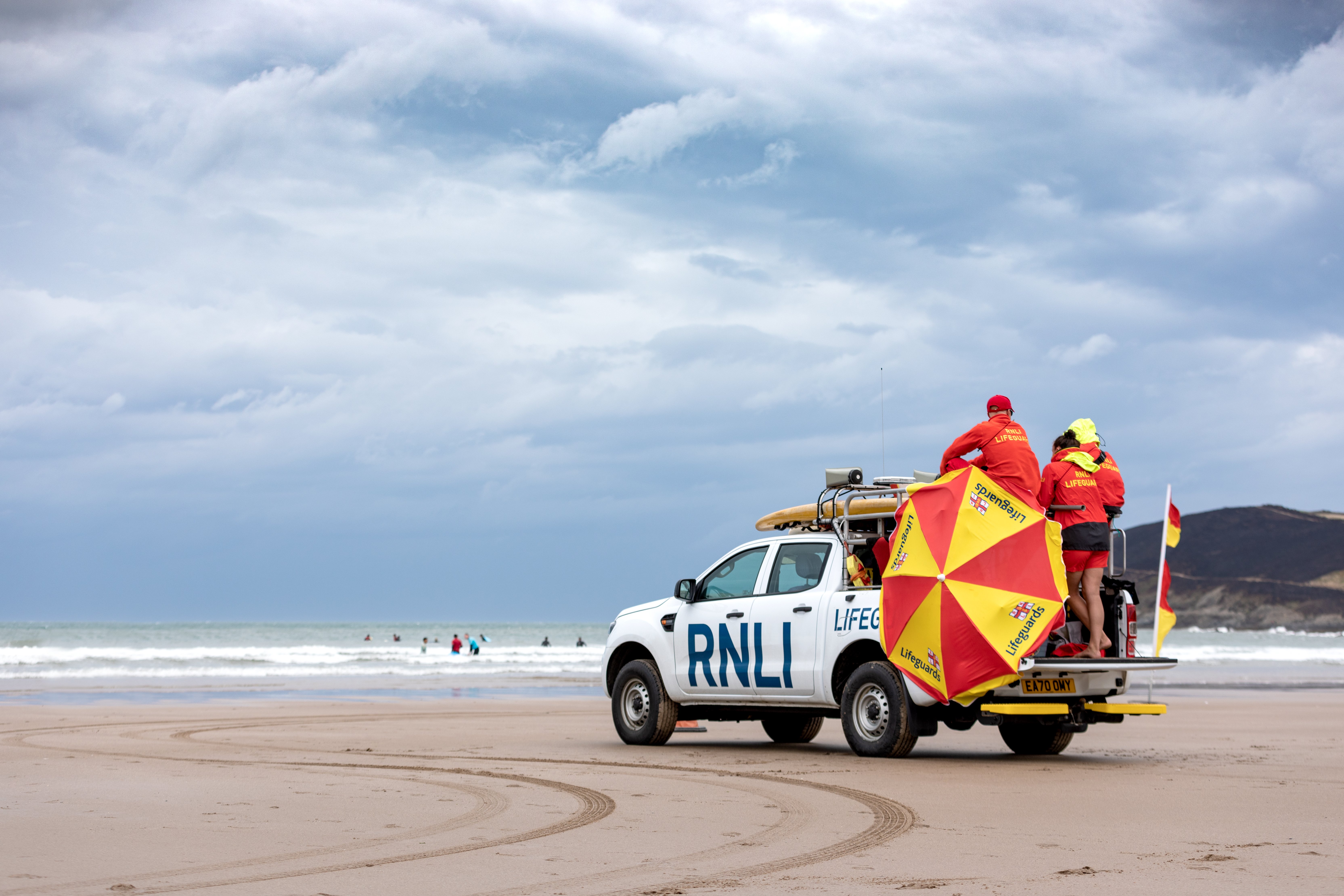 A white RNLI truck patrolling a sandy beach carrying lifeguards wearing red and yellow clothes