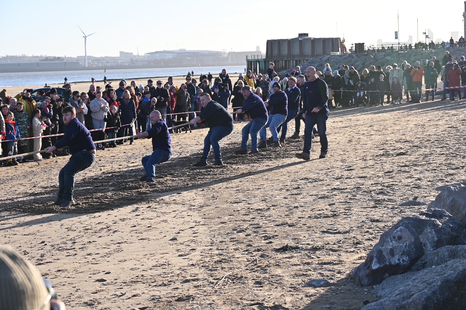 New Brighton beach. A tug of war team from New Brighton RNLI all pull together on a rope. Picture captured mid-pull. In the background crowds watch on.