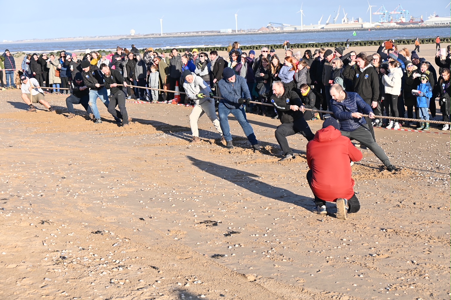 A beach scene. The tug of war team from Mersey Divers are captured, mid-pull, whilst crowds watch on in the background.