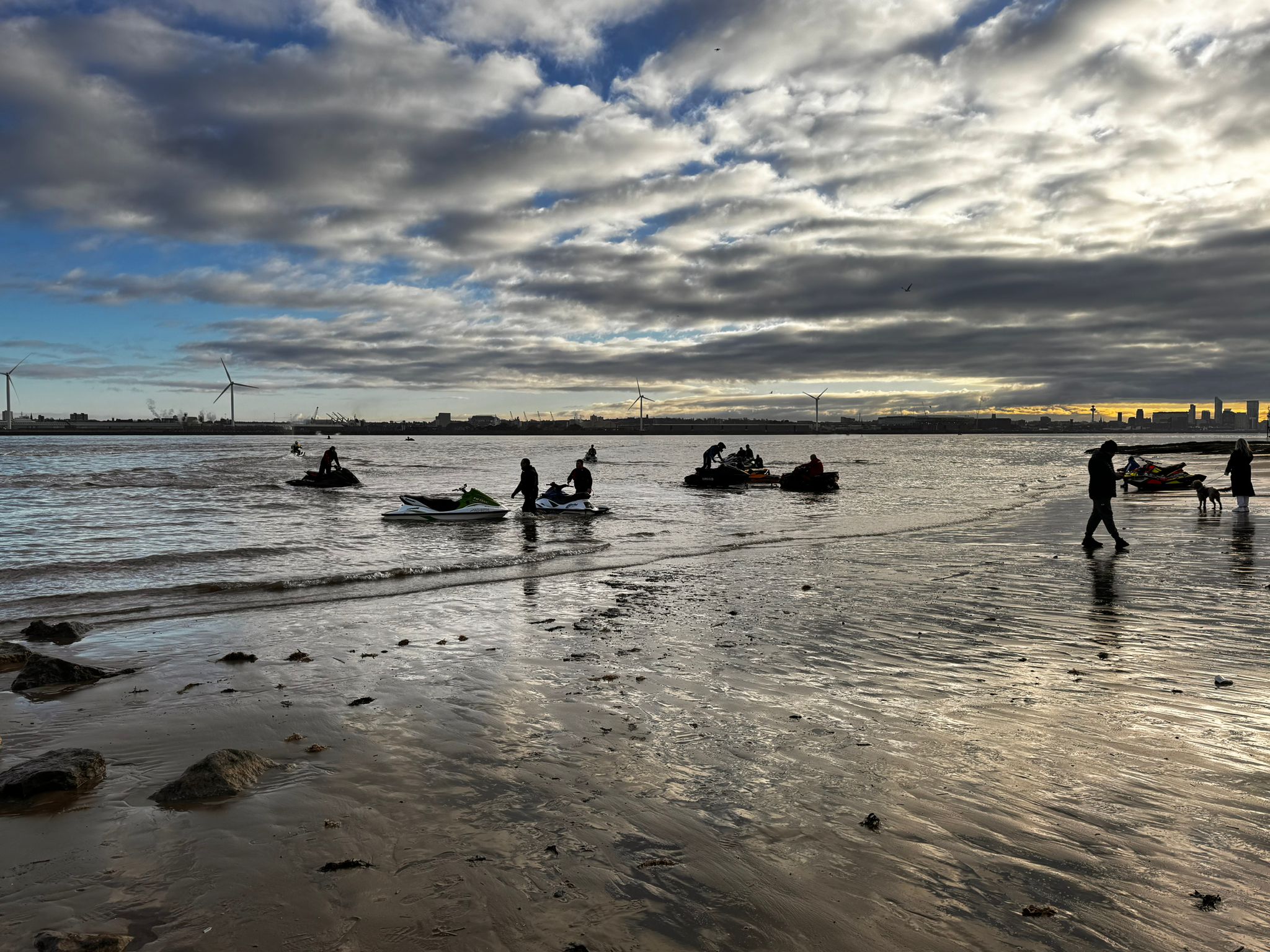 A late afternoon beach and river scene, with dramatic cloudy sky. Multiple jet skis can be seen in the shallow waters of the Mersey, with people embarking or disembarking.