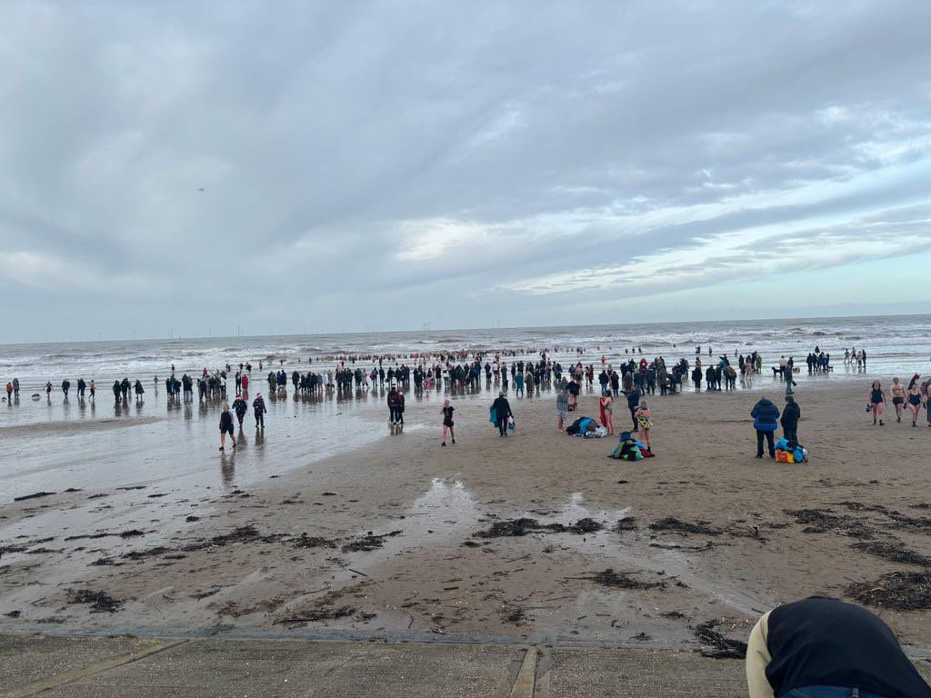 A beach scene. Crowds of swimmers can be seen on the sand, the water's edge and in the water.