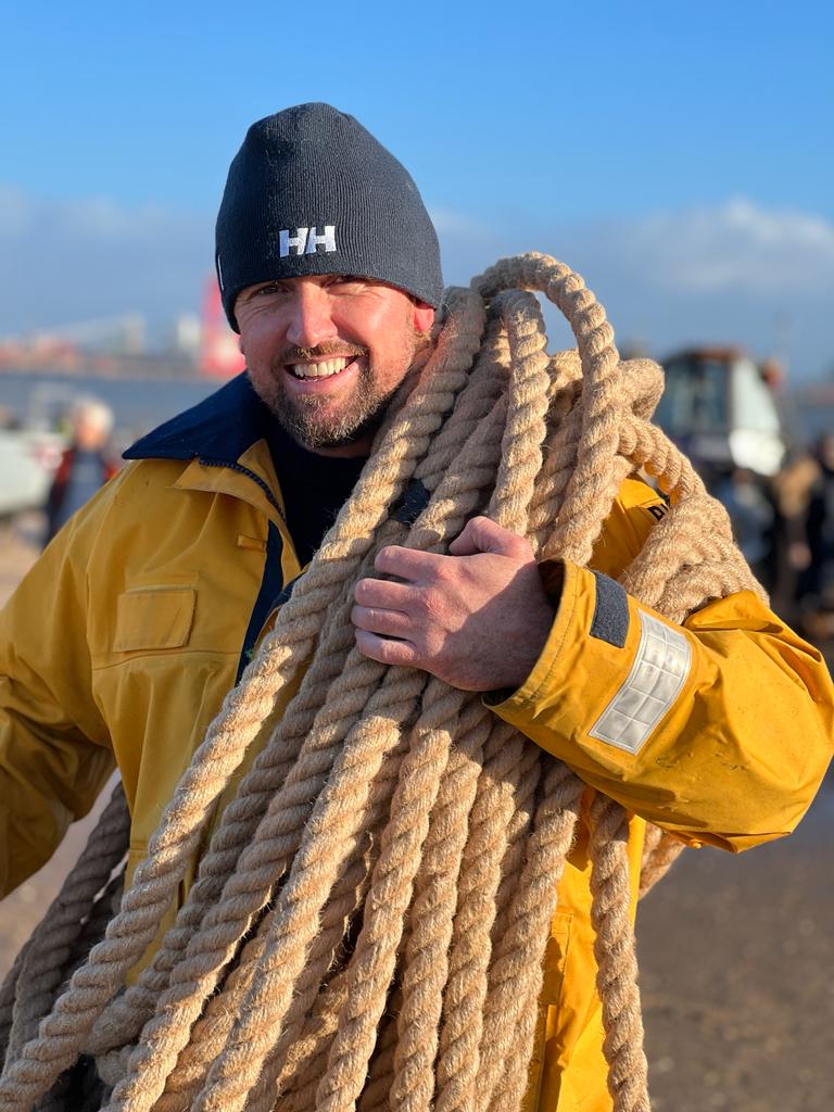 A New Brighton RNLI crew member is framed fully, midshot, in beanie hat and yellow waterproof jacket, tug of war rope wrapped around his shoulders, transporting it back to be stored for another year.