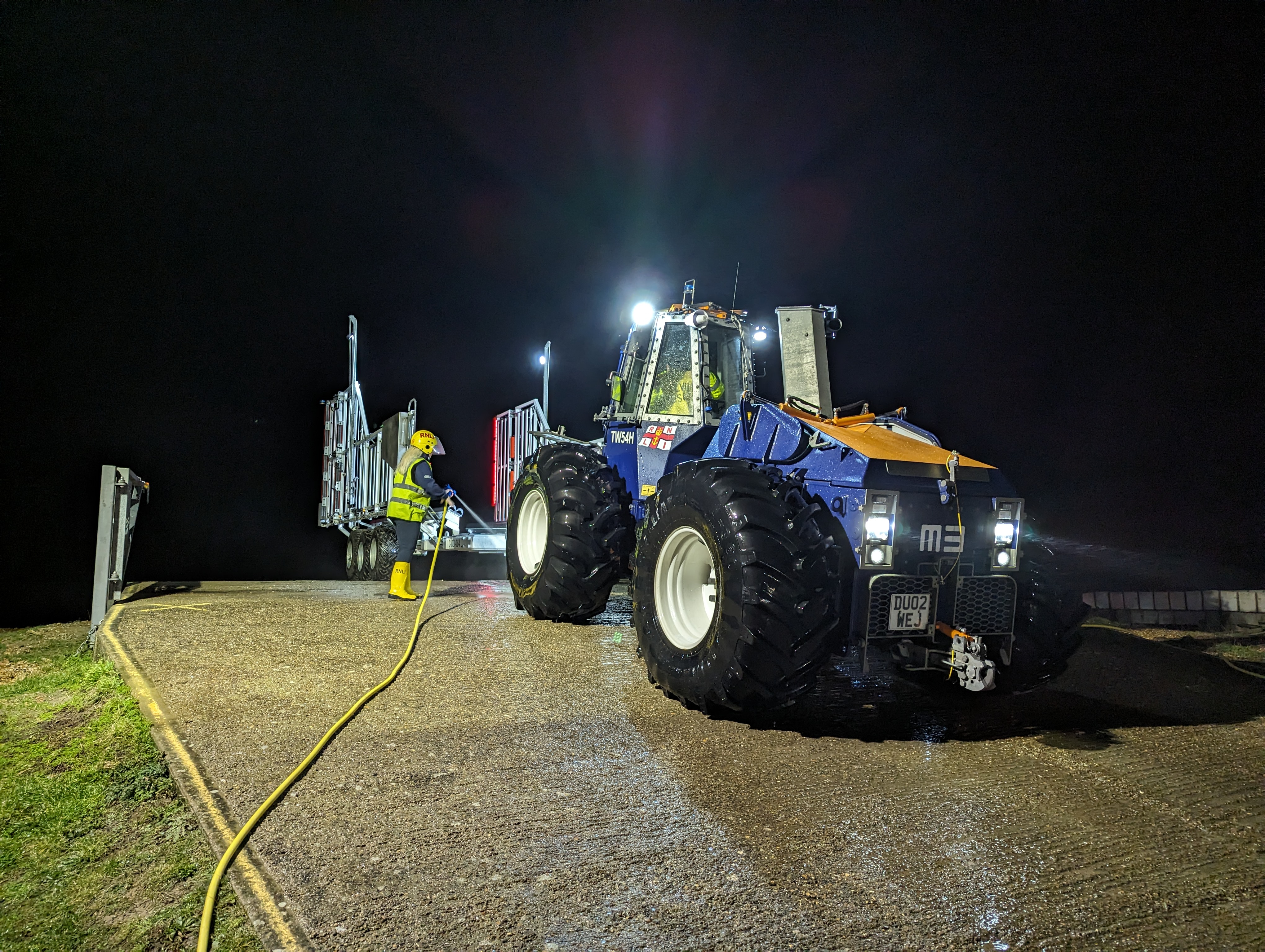 A member of shore crew in high Vis jacket sprays down the boat and tractor after a training launch on a dark and cold night