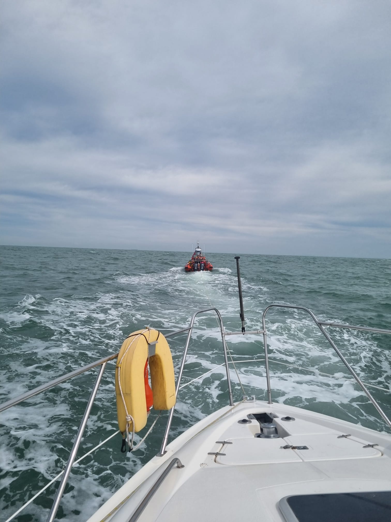 An RNLI orange lifeboat, the Jean McIvor, is running through the seas ahead of a motor yacht with a tow in place