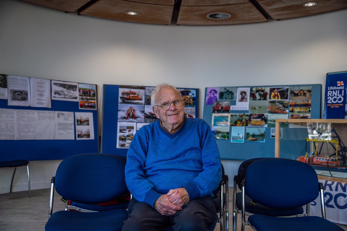 Man in blue jumper sitting on blue chair with photos on boards behind him 