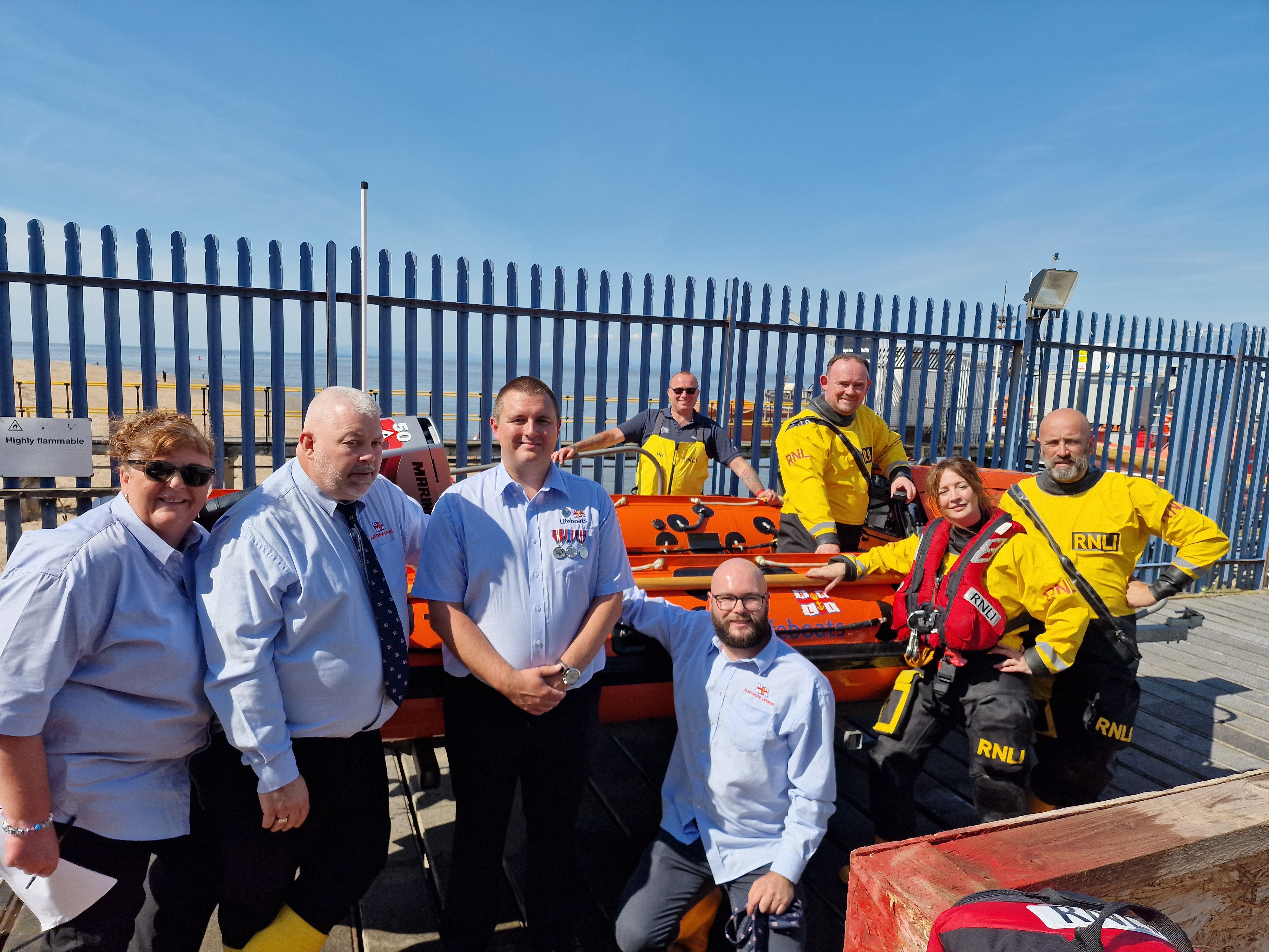 A group of crew in blue shirts, some with ties, and the crew that launched in their black &amp; yellow RNLI drysuits, with the D Class lifeboat Harbet