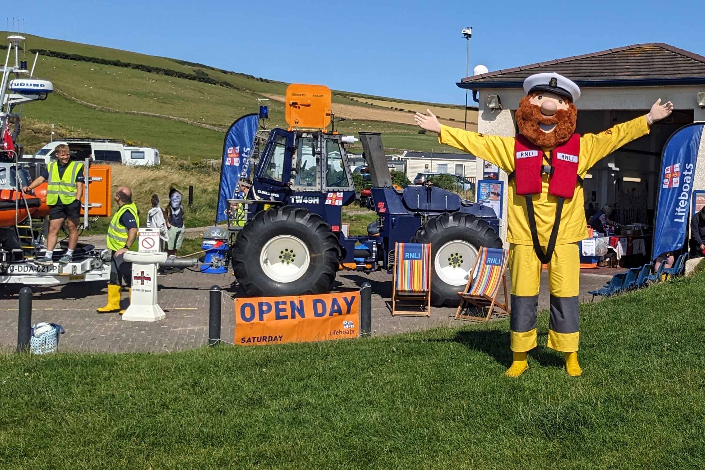 Stormy Stan the RNLI mascot has his arms open to welcome visitors standing in front of the tractor and lifeboat station