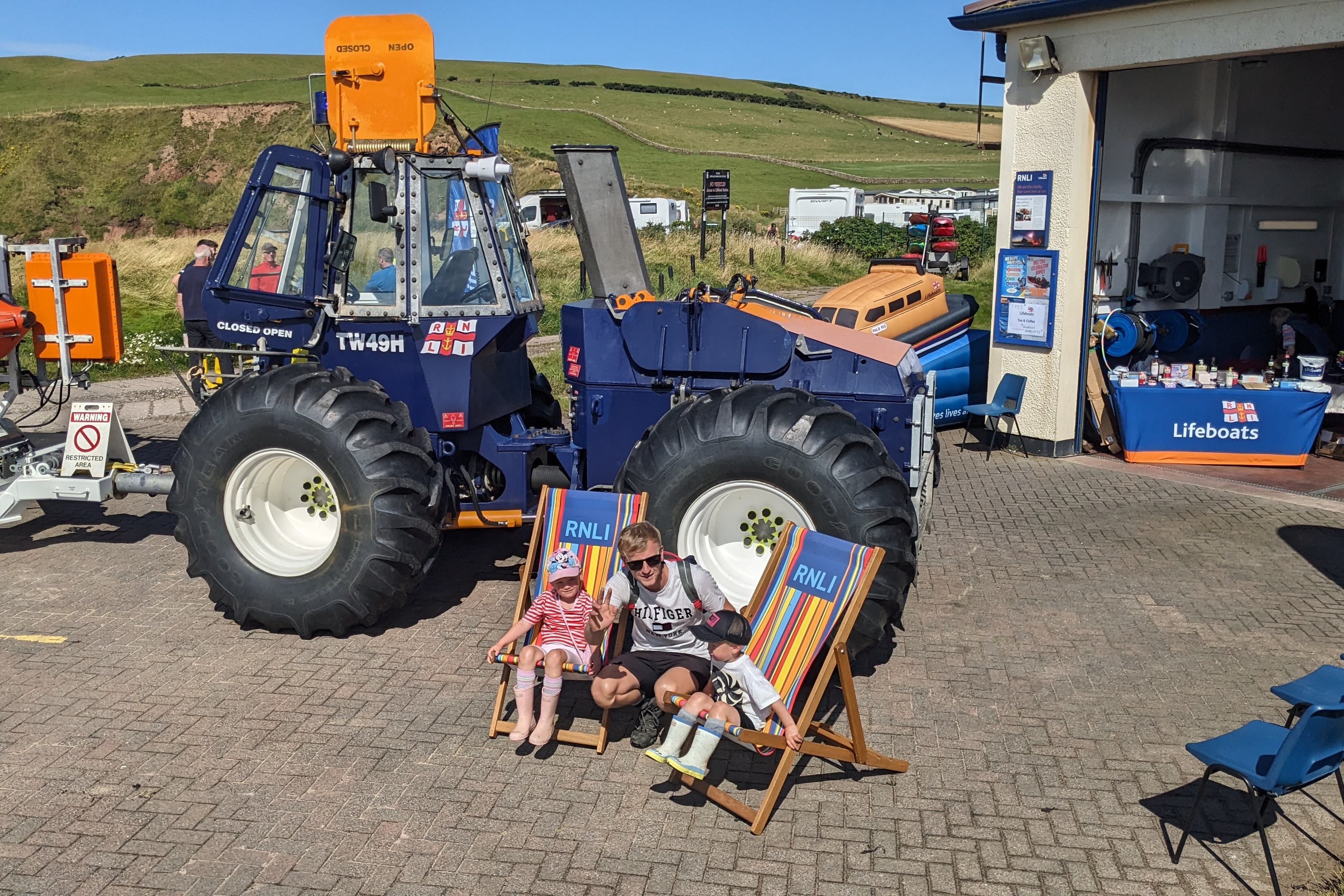 A father and his two young children take a rest in the large RNLI deckchairs beside the RNLI tractor