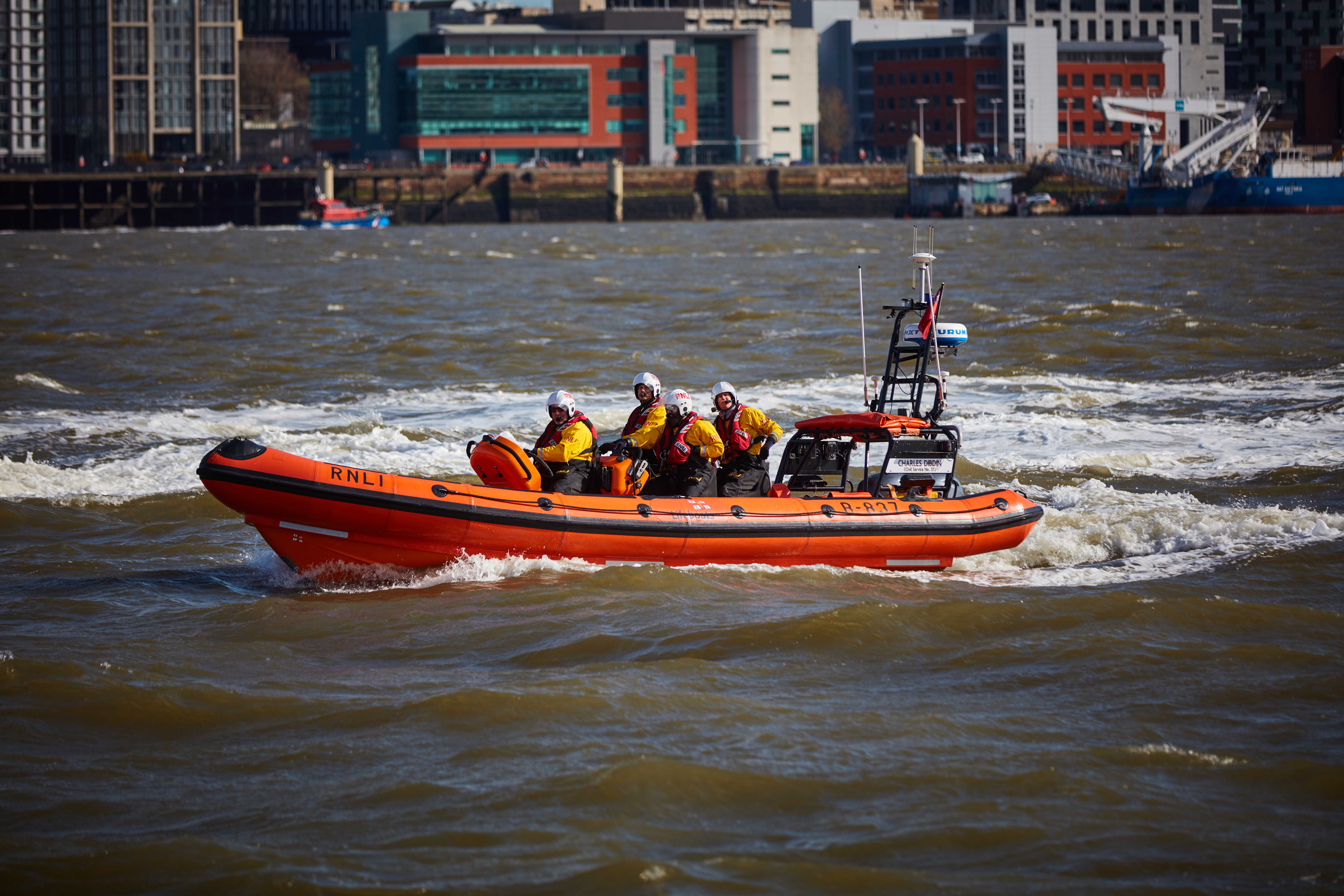 New Brighton's Atlantic 85 lifeboat, with volunteer crew on board, is the subject of the image. The River Mersey is in the foreground and background.
