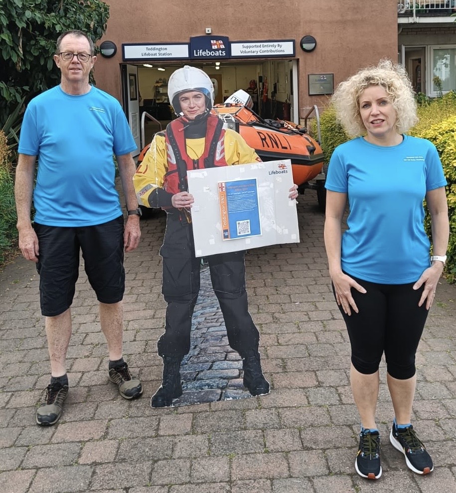 Ian and Jan in front of the lifeboat station wearing their new running shirts which say Teddington RNLI 200 km Relay Challenge on the front with a large RNLI flag on the back.