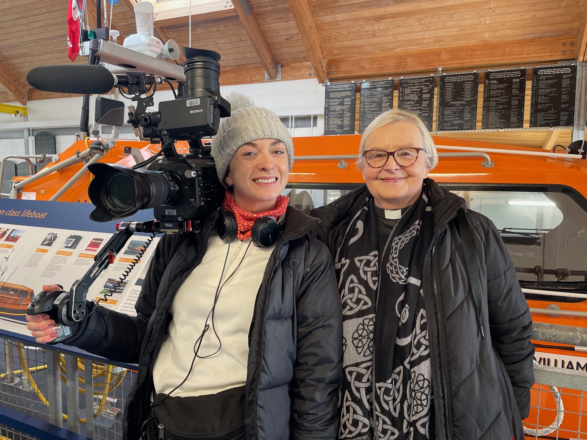 Dark haired woman with a filming camera with a woman wearing a priest's dog collar