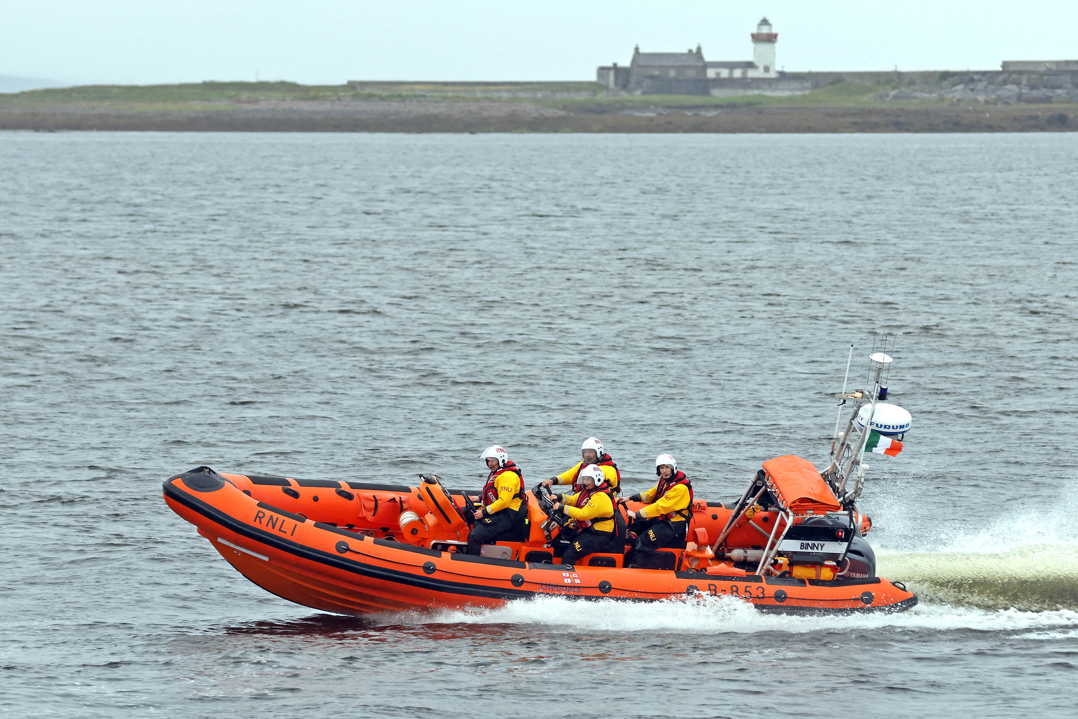 Four crew on board an Atlantic 85 lifeboat at sea moving from right to left with Mutton Island and Lighthouse in the distance behind