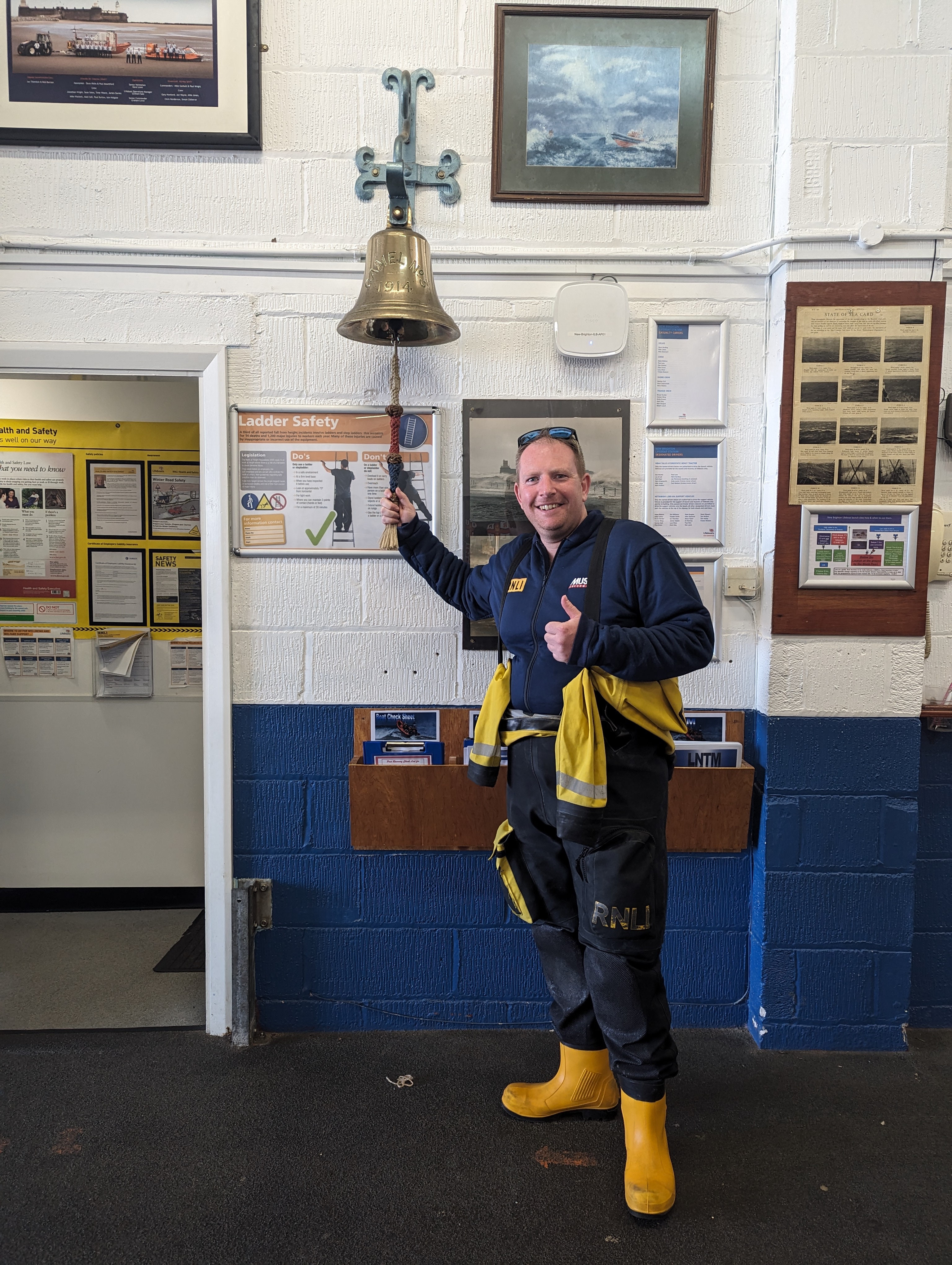Adam Bidston, newest helm at New Brighton RNLI, is seen posing, thumbs up and smiling, ringing the bell at New Brighton boathouse.