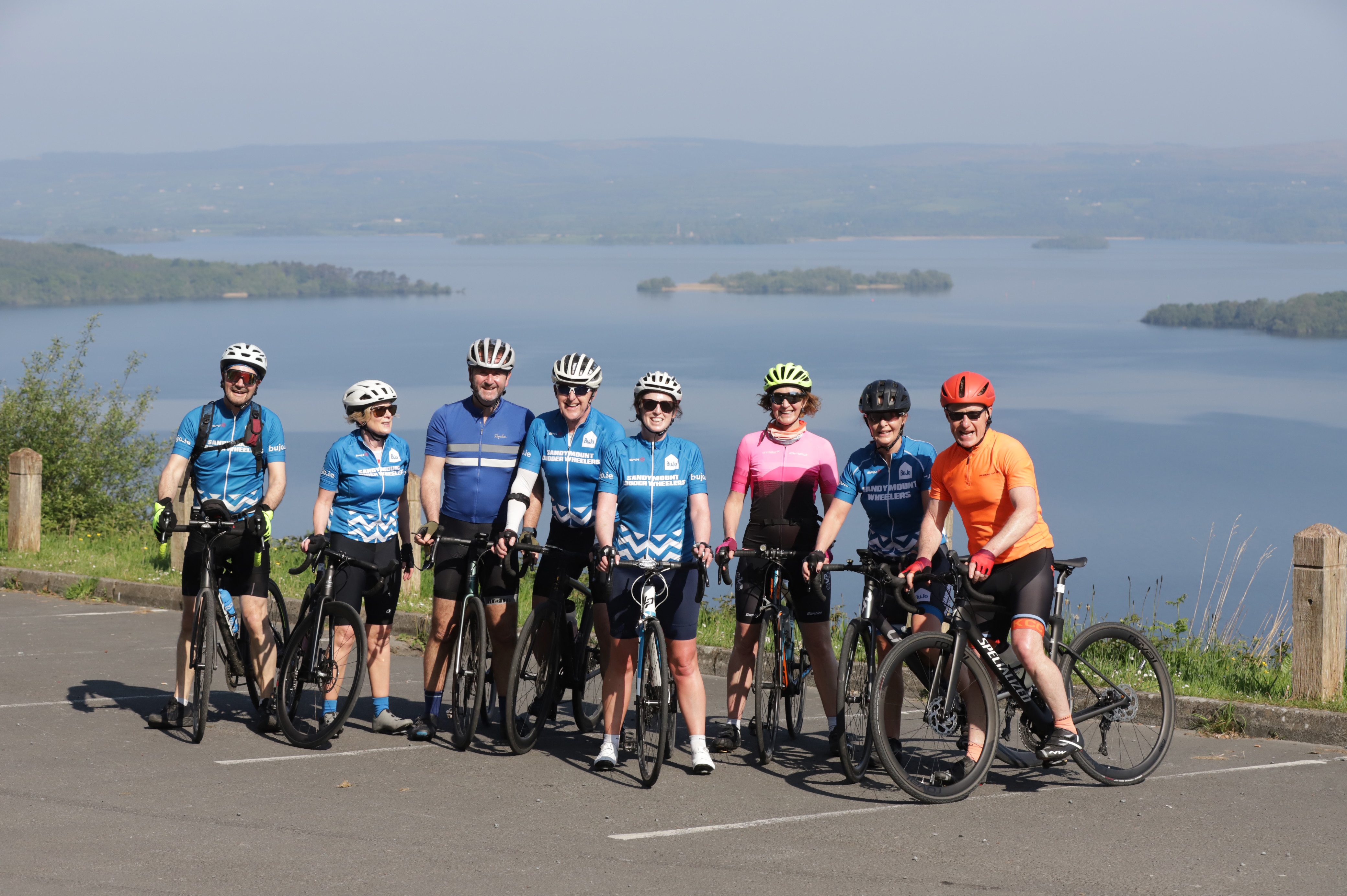 8 cyclists pose at the lookout with a panorama below them of Lough Derg