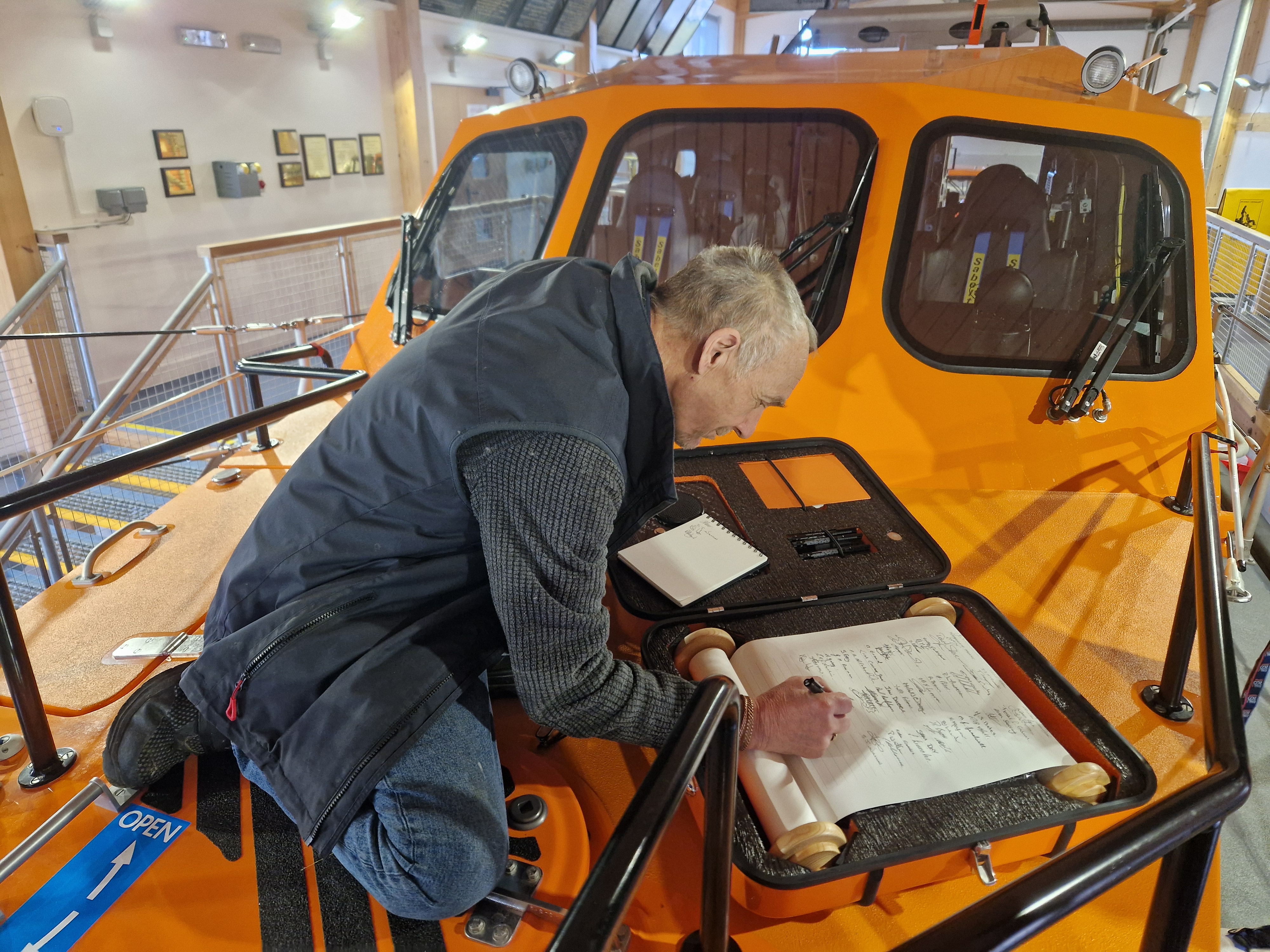 Crew member signing the scroll with pen in hand and lifeboat in background
