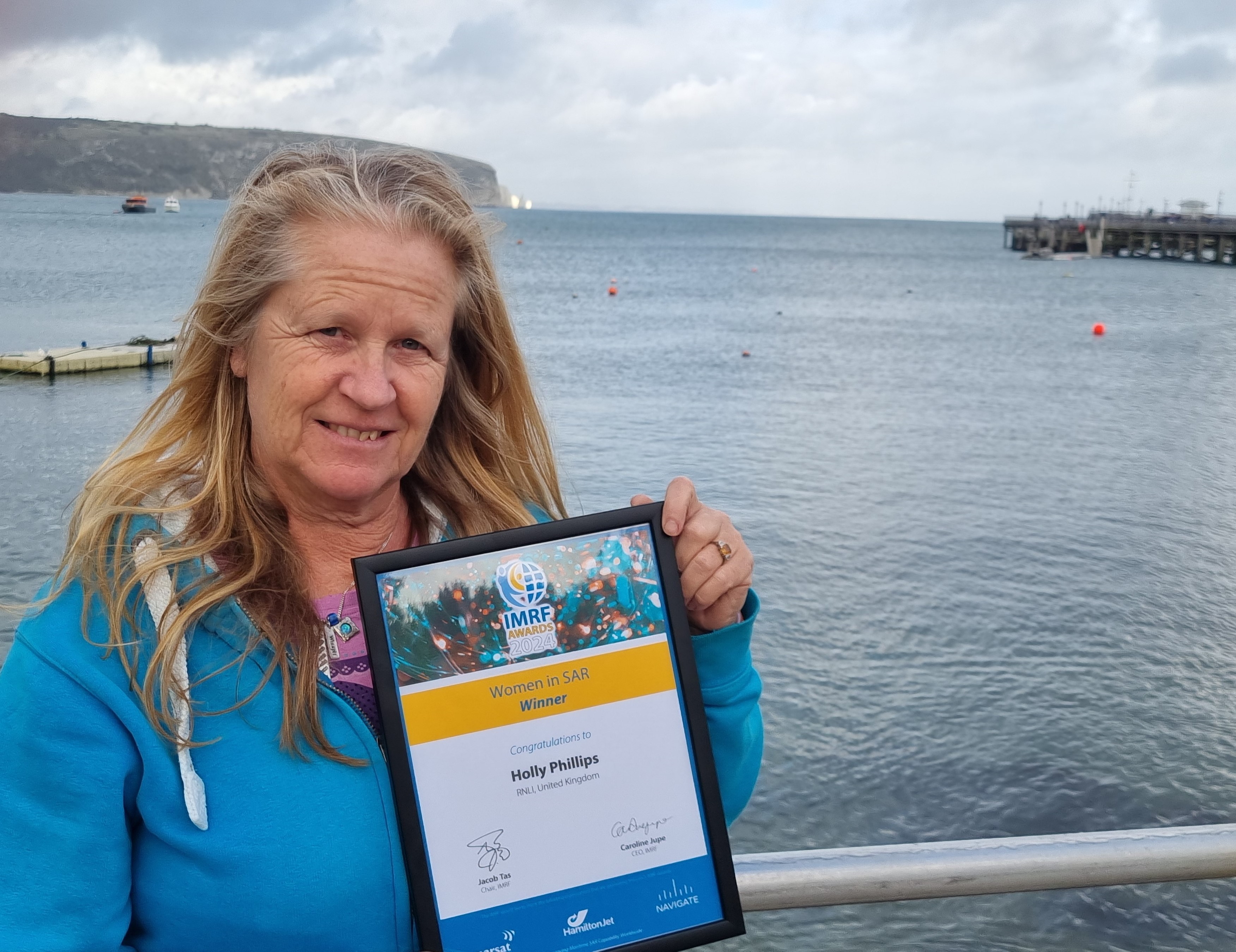 Woman holds framed award in front of sea backdrop.
