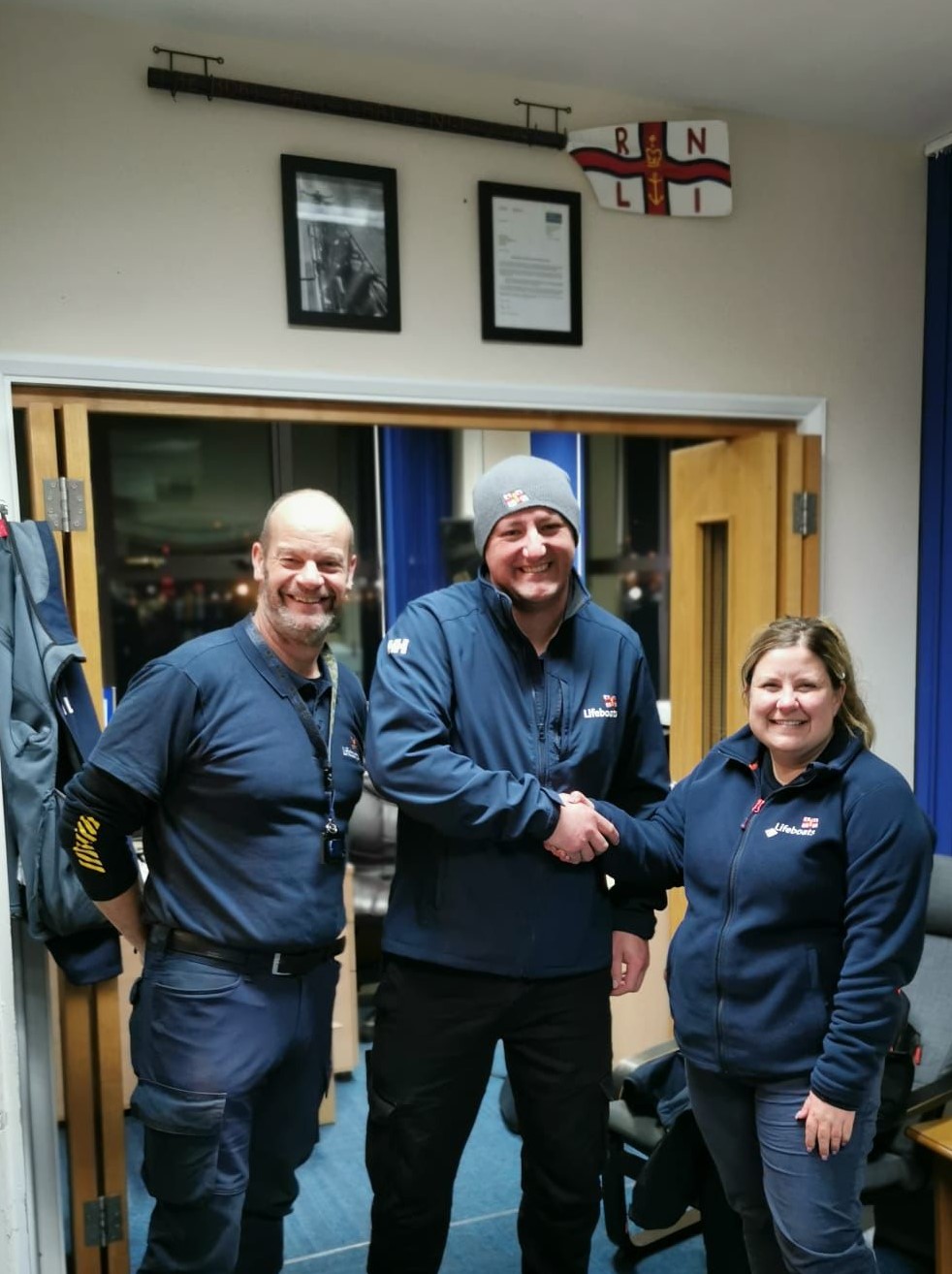 Three people in navy RNLI branded clothing stand indoors smiling at the camera. The man in the centre shakes the hand of the woman on the right.
