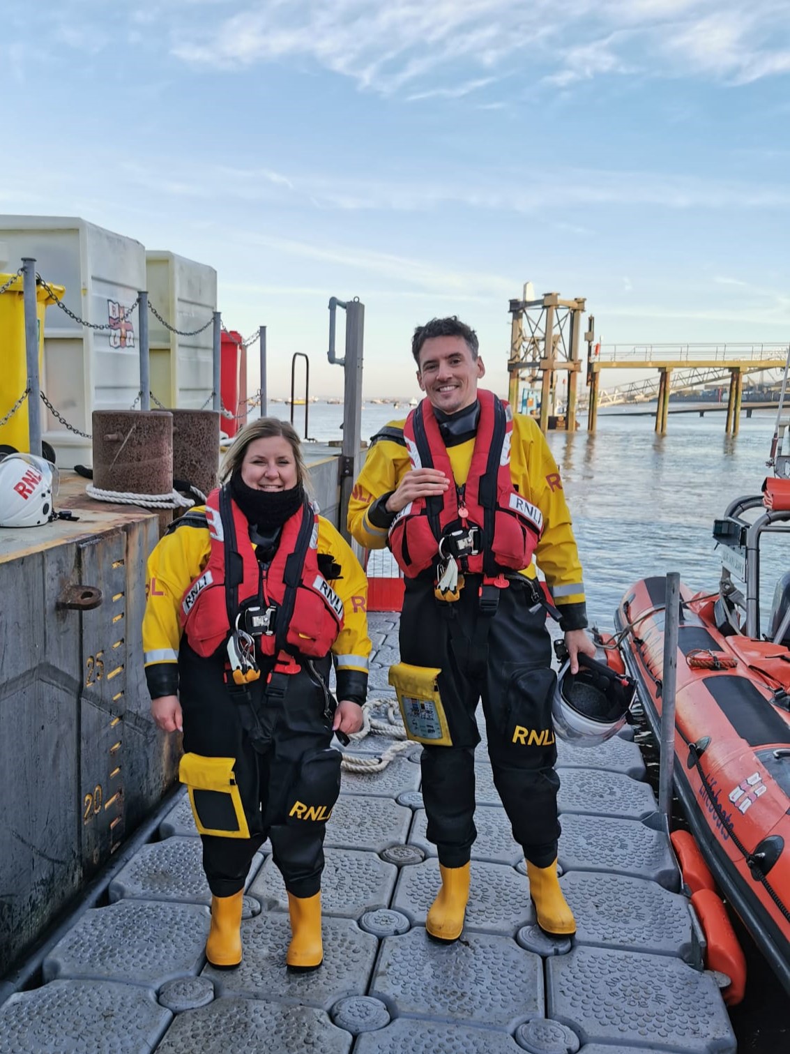 A man in a yellow drysuit, and red lifejacket stands between a woman in a yellow drysuit, and red lifejacket and an orange Atlantic 85 lifeboat