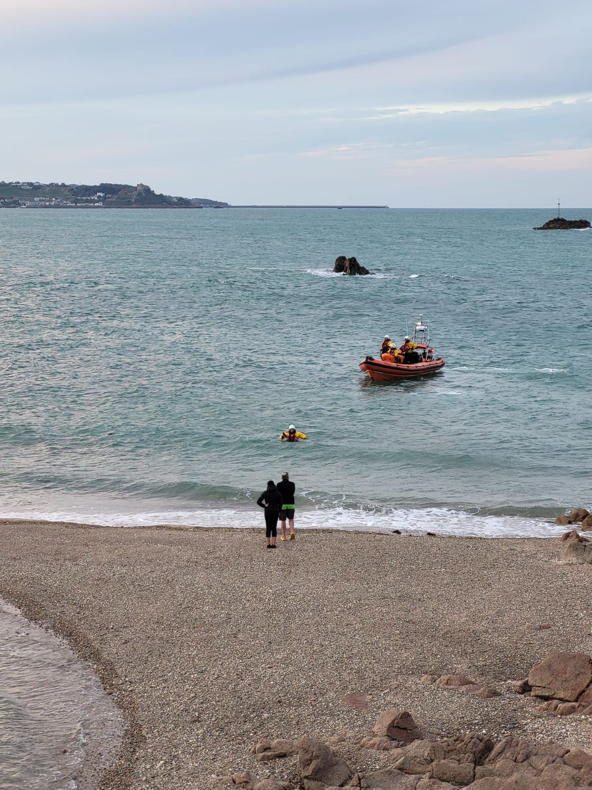 A crew member makes their way ashore from the lifeboat towards two members of the public standing on teh shore. The crew member is chest deep in the water as the lifeboat stands off.