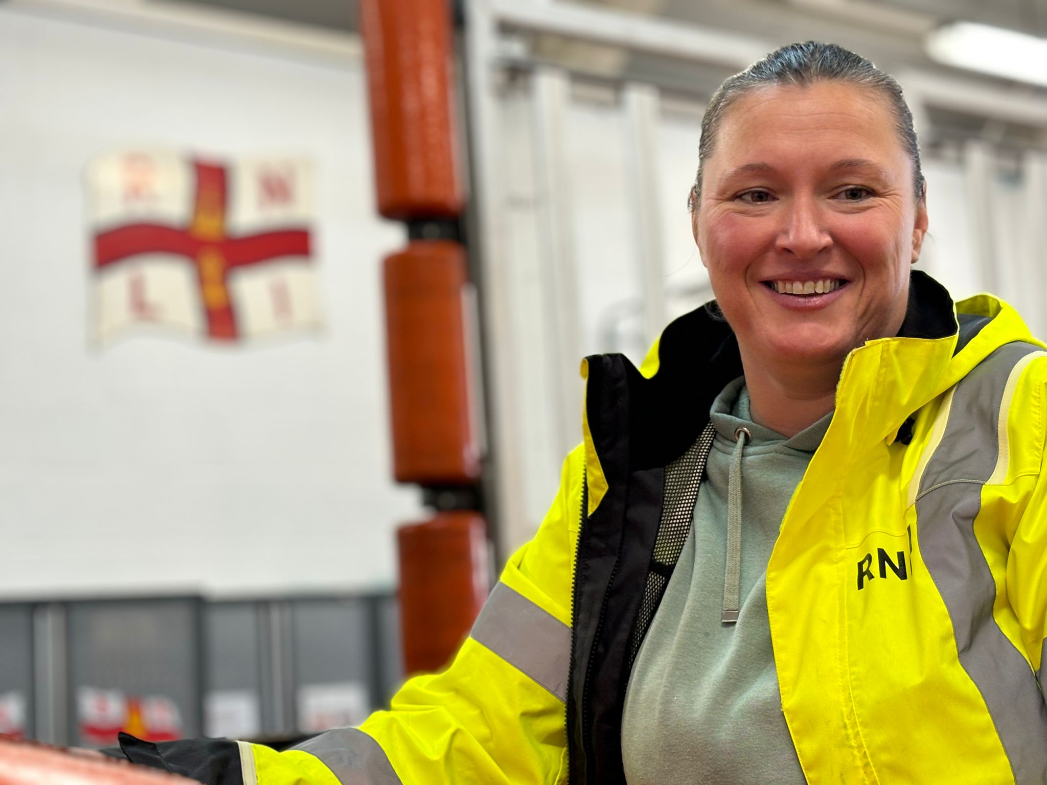 Fliss is in the foreground in RNLI high vis jacket, smiling at the camera. In soft focus behind can be seen the RNLI flag and the carriage for the boat.