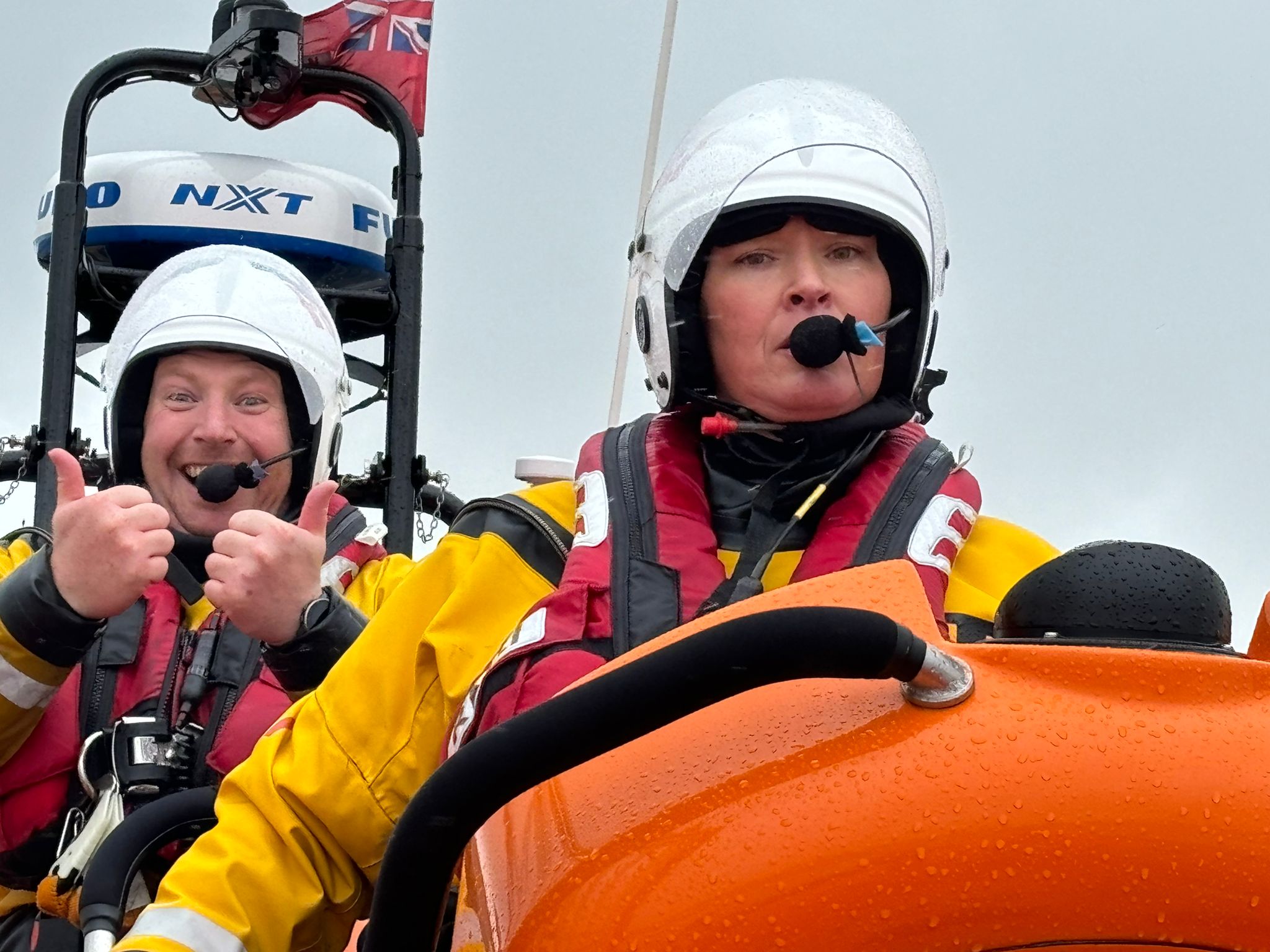 Fliss is on board the atlantic-85 lifeboat in the foreground, bringing it in to recover. Over her right shoulder a crew member can be seen smiling and giving a big thumbs up to the camera, out of sight of Fliss.