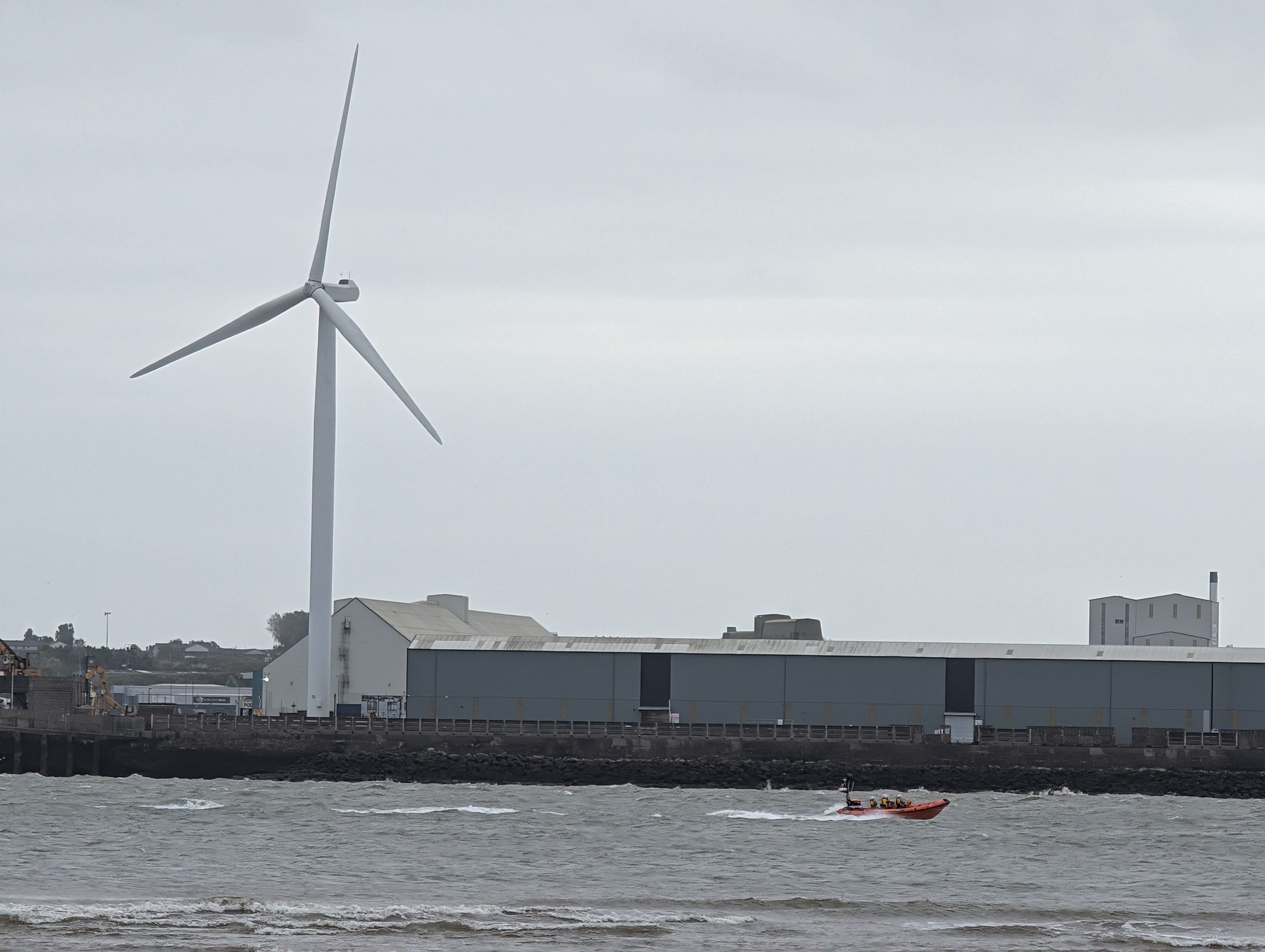 A photo from the shoreside shows the Atlantic-85 speeding past from left to right. In the background can be seen some warehouses and a wind turbine.