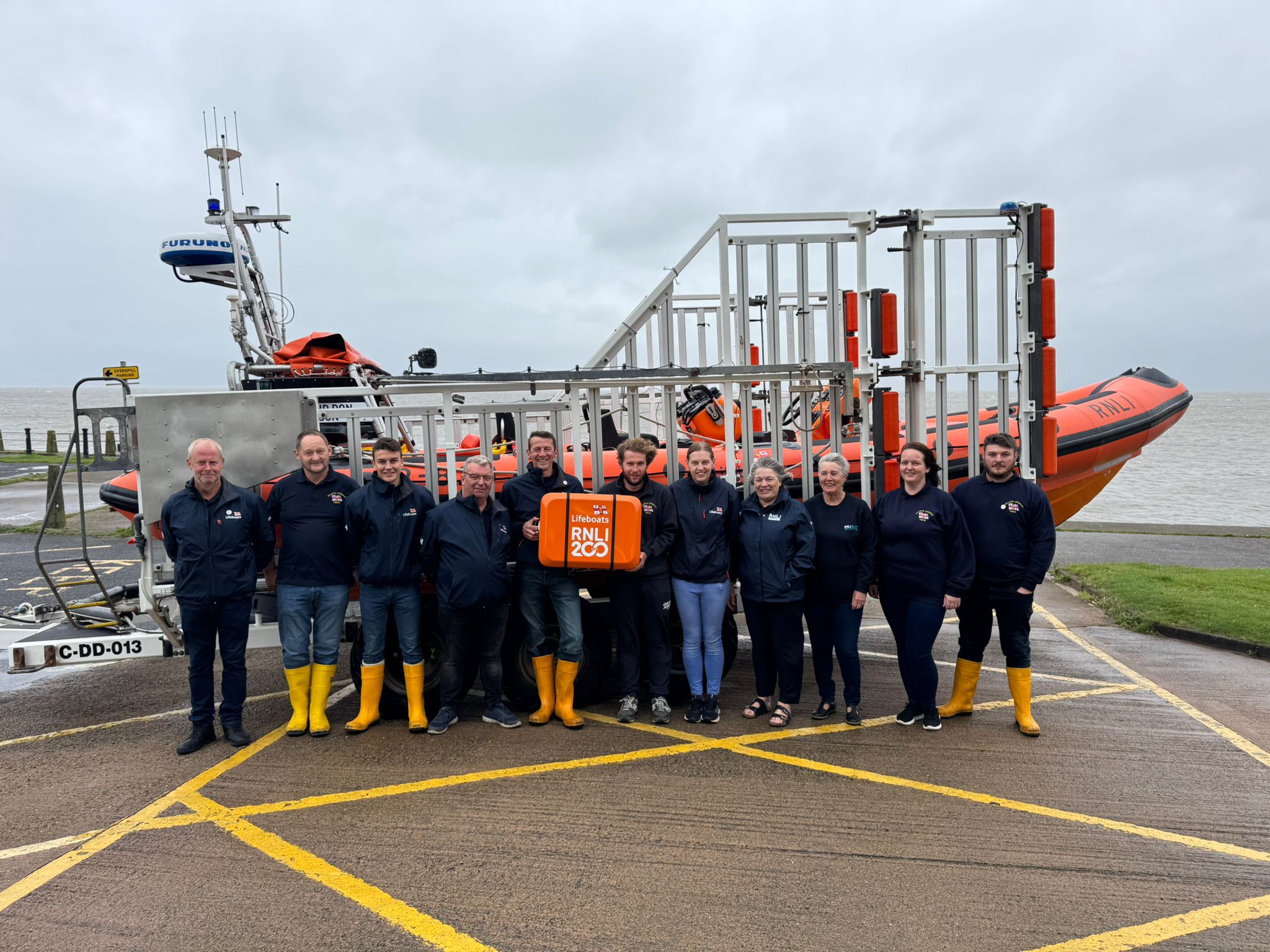 Crew standing in front of orange lifeboat holding orange scroll box 