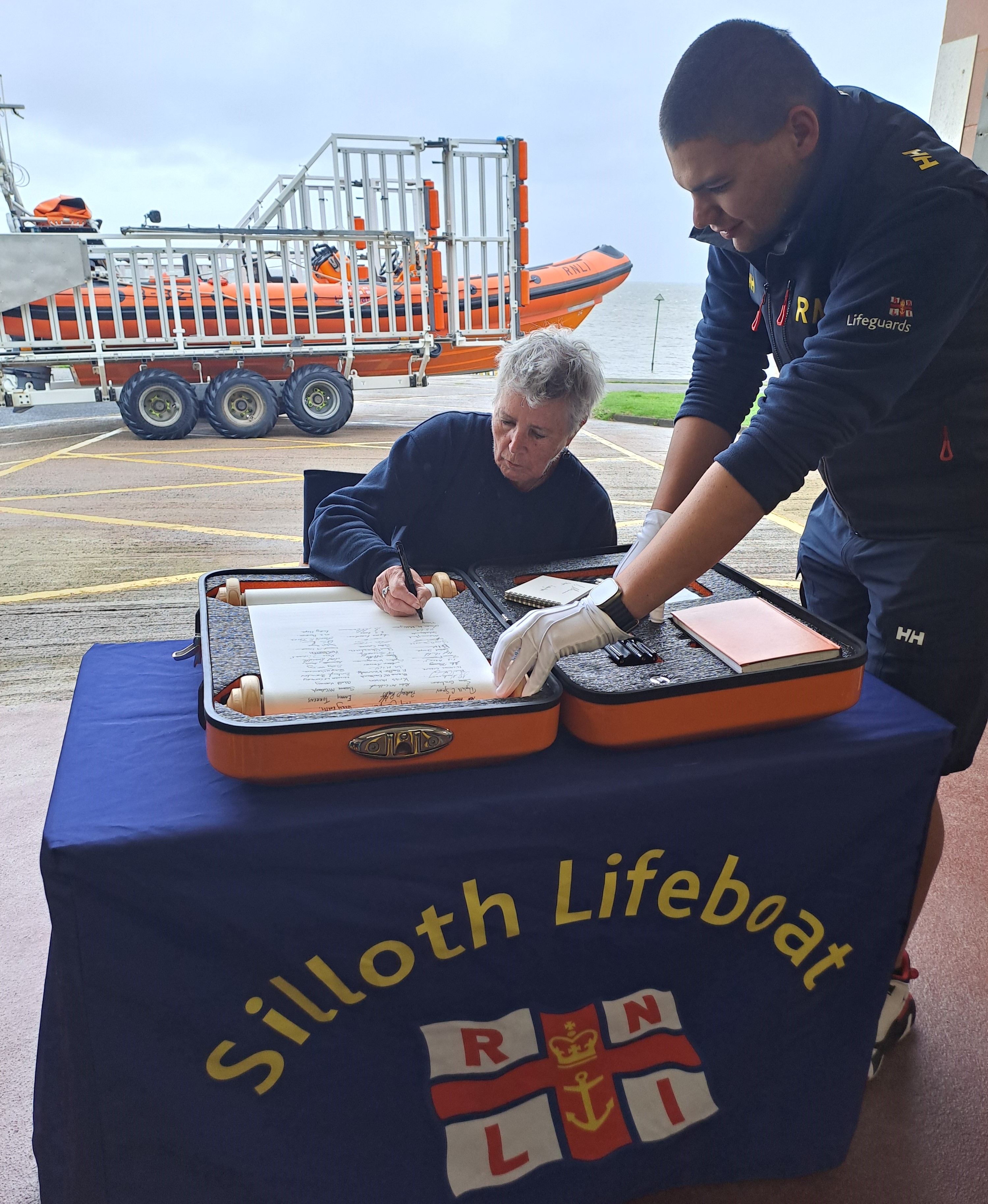 Female sitting on chair behind table signing scroll with orange lifeboat in the background