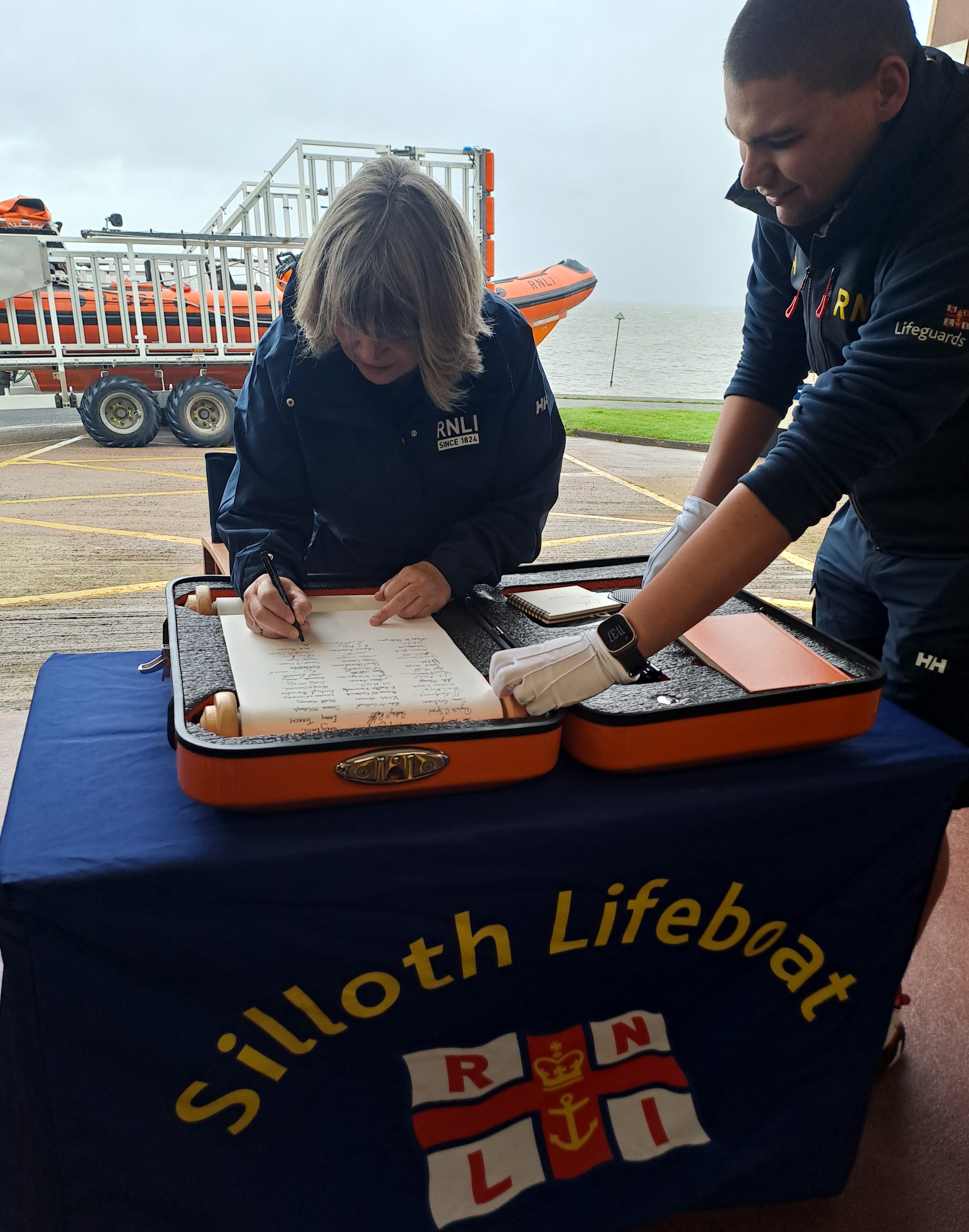 Female standing behind table signing scroll with orange lifeboat in the background