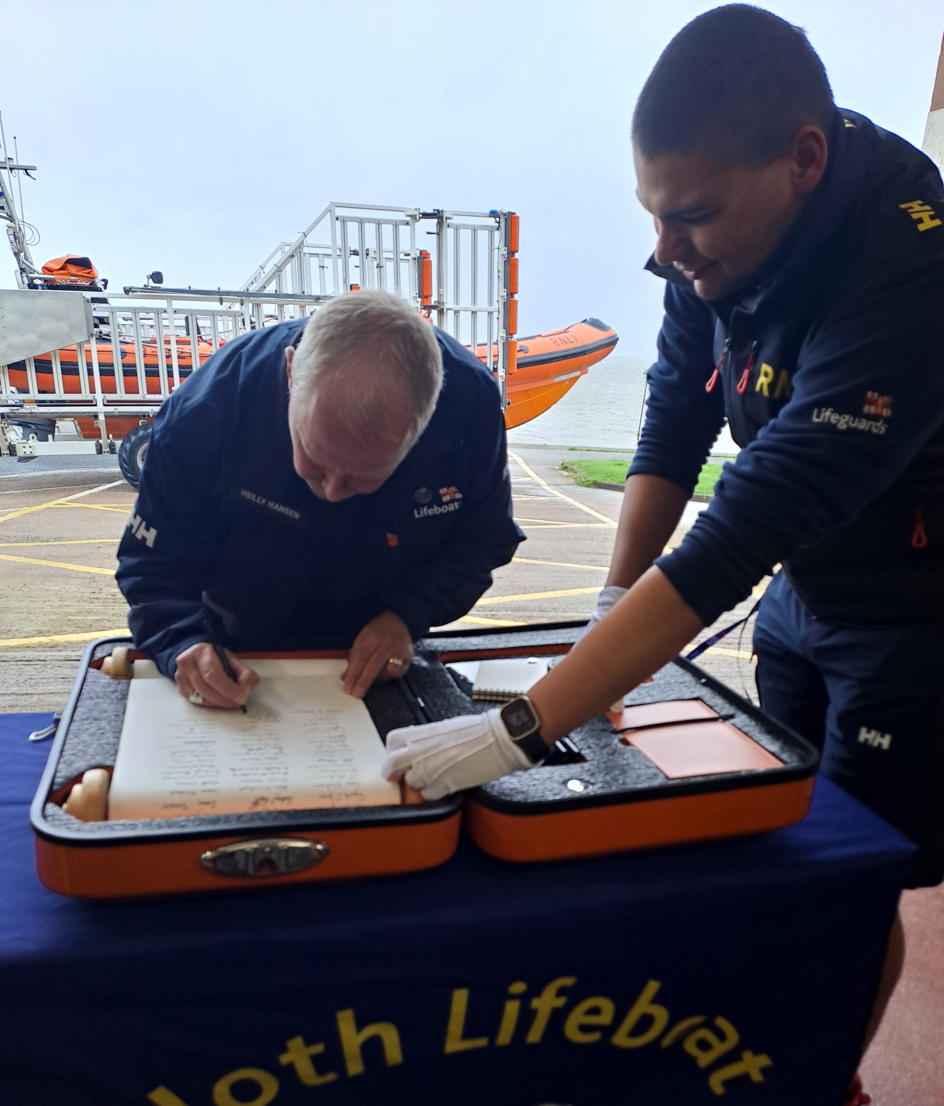 Male sitting on chair behind table signing scroll with orange lifeboat in the background