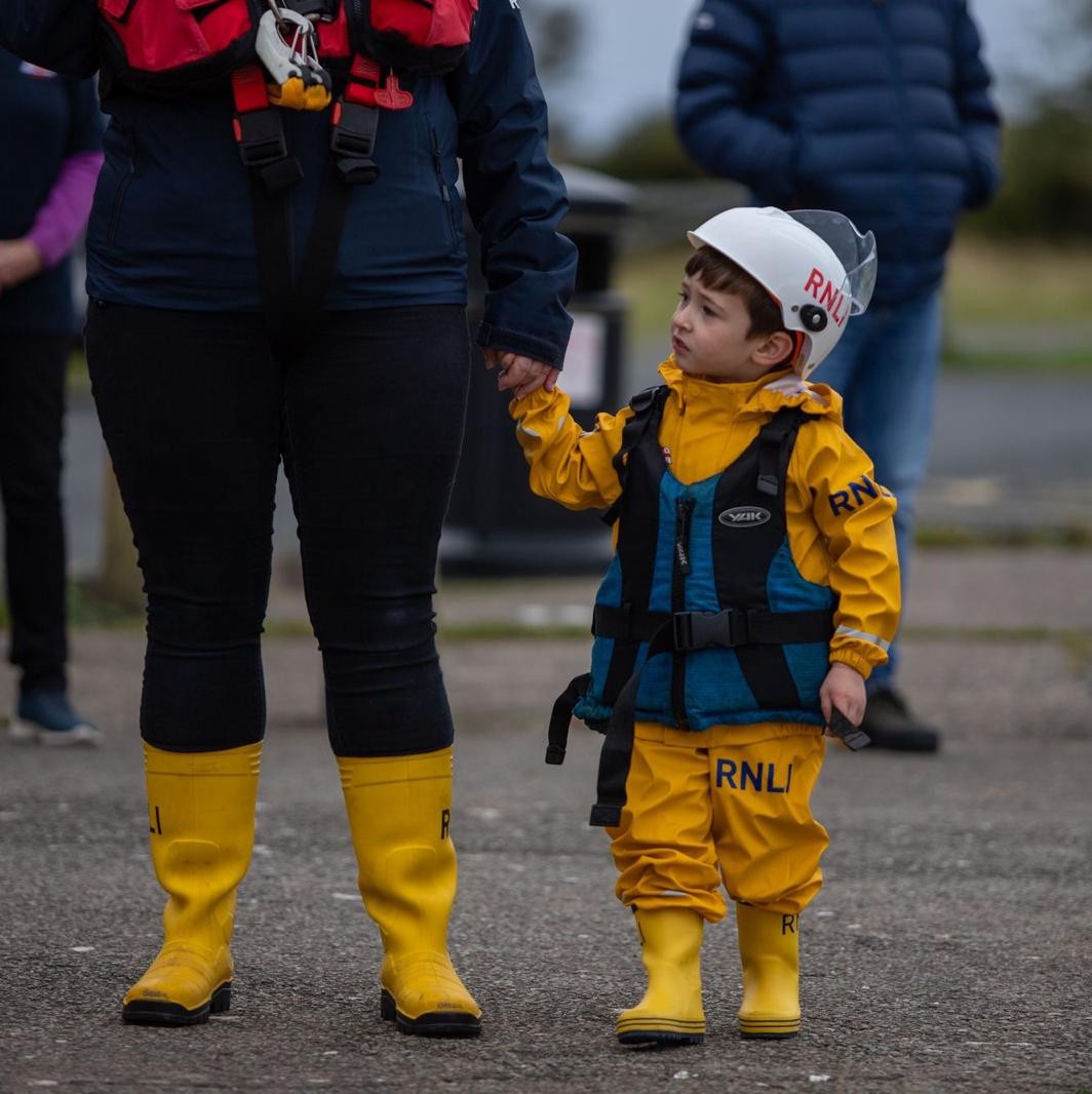Child dressed in yellow clothing and yellow wellie wearing blue life vest holding mother's hand looking to lifeboat beside them