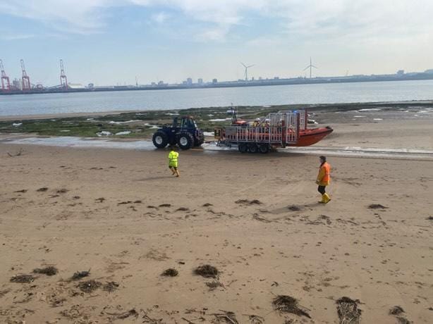 New Brighton beach, with shore crew visible in hi-vis gear escorting the Bendy tractor, carriage and lifeboat to the water's edge. In the background the Mersey can be seen.