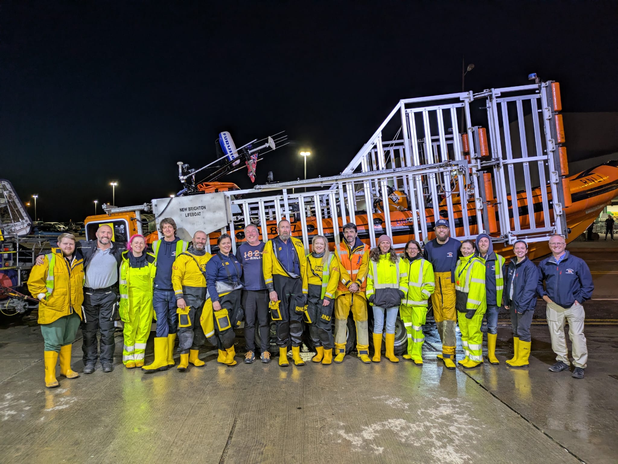 Night time, a large group of New Brighton RNLI volunteers pose in front of their lifeboat, held on the carriage, outside the boathouse.
