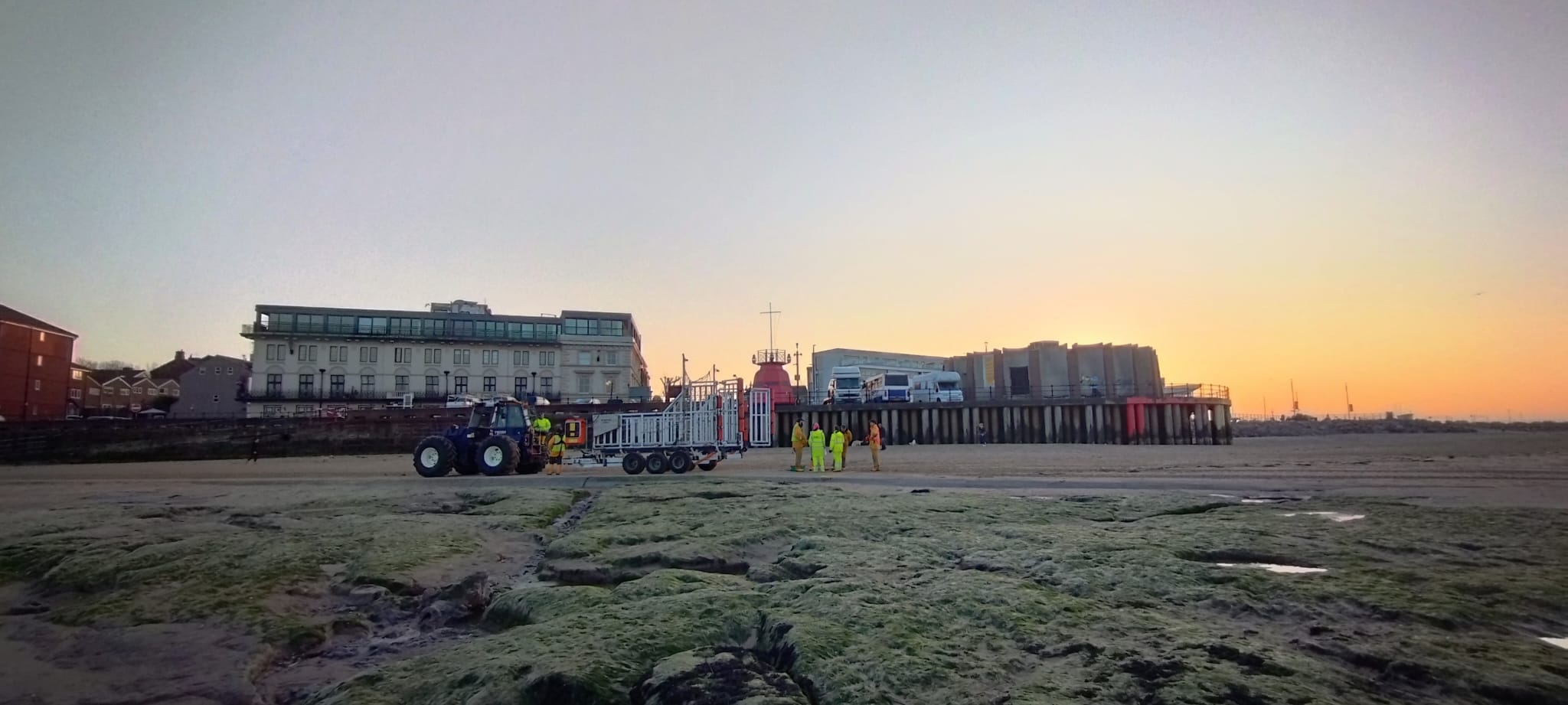 A wide angle shot from a distance. Sunset, with beautiful golden colours in the sky. Silhouetted against the beach and the old buildings beyond, New Brighton RNLI's tractor, carriage and lifeboat can be seen moving up the beach, with shore crew either side, escorting.