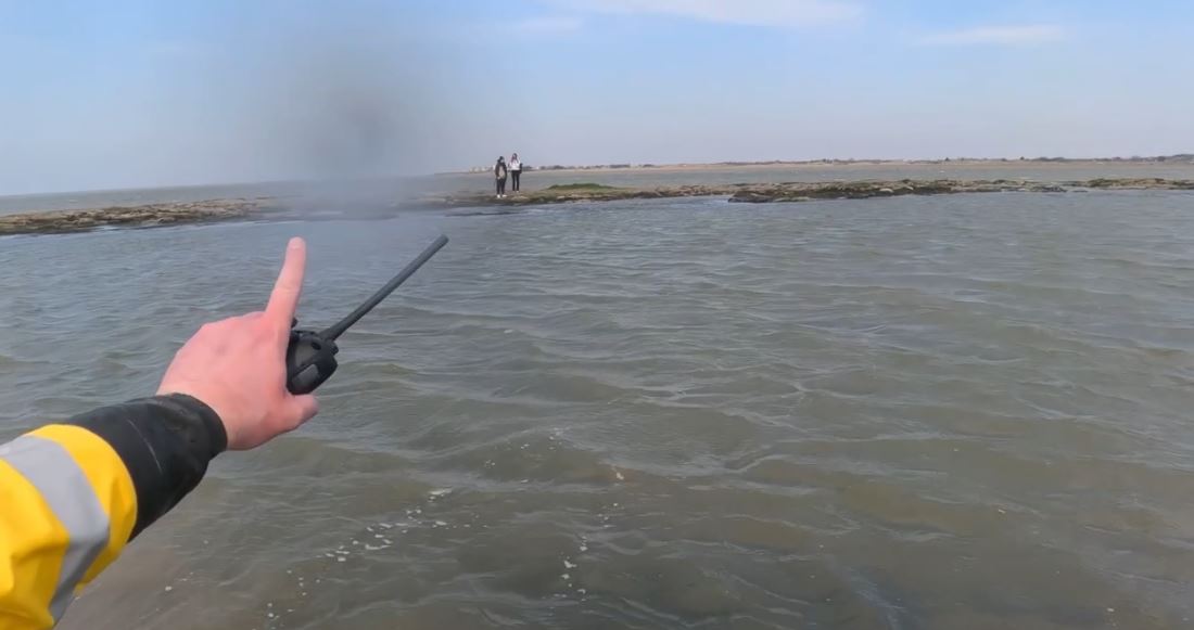 An arm wearing a yellow drysuit with black cuff points to two people in the distance surrounded by the sea.