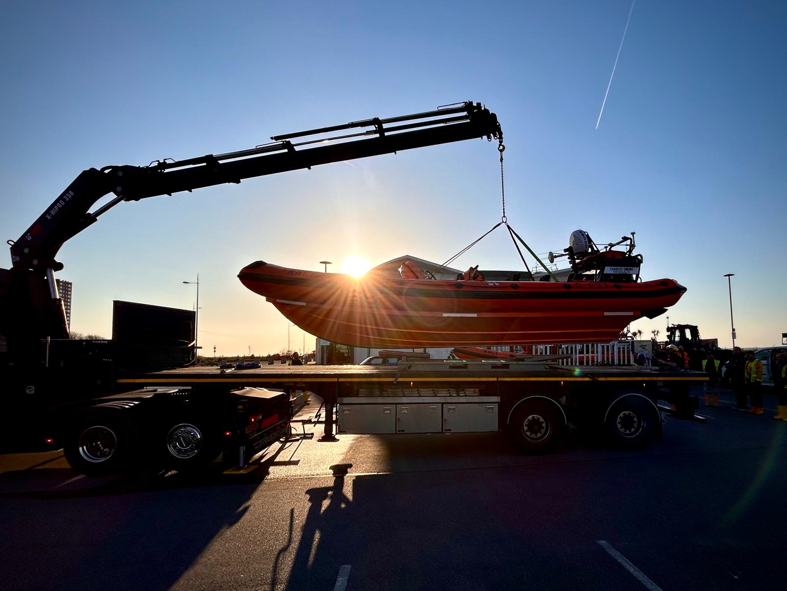 A beautiful sunset sky in New Brighton. Silhouetted against the sunset the lifeboat can be seen, attached to a crane arm, being lifted from the back of a flat bed truck.
