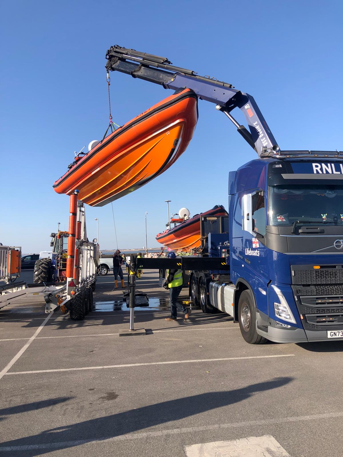 The B-class lifeboat can be seen suspended in mid-air, lifted by crane arm, as it is moved out of the now empty carriage to the left of the frame, to the flat bed truck on the right.
