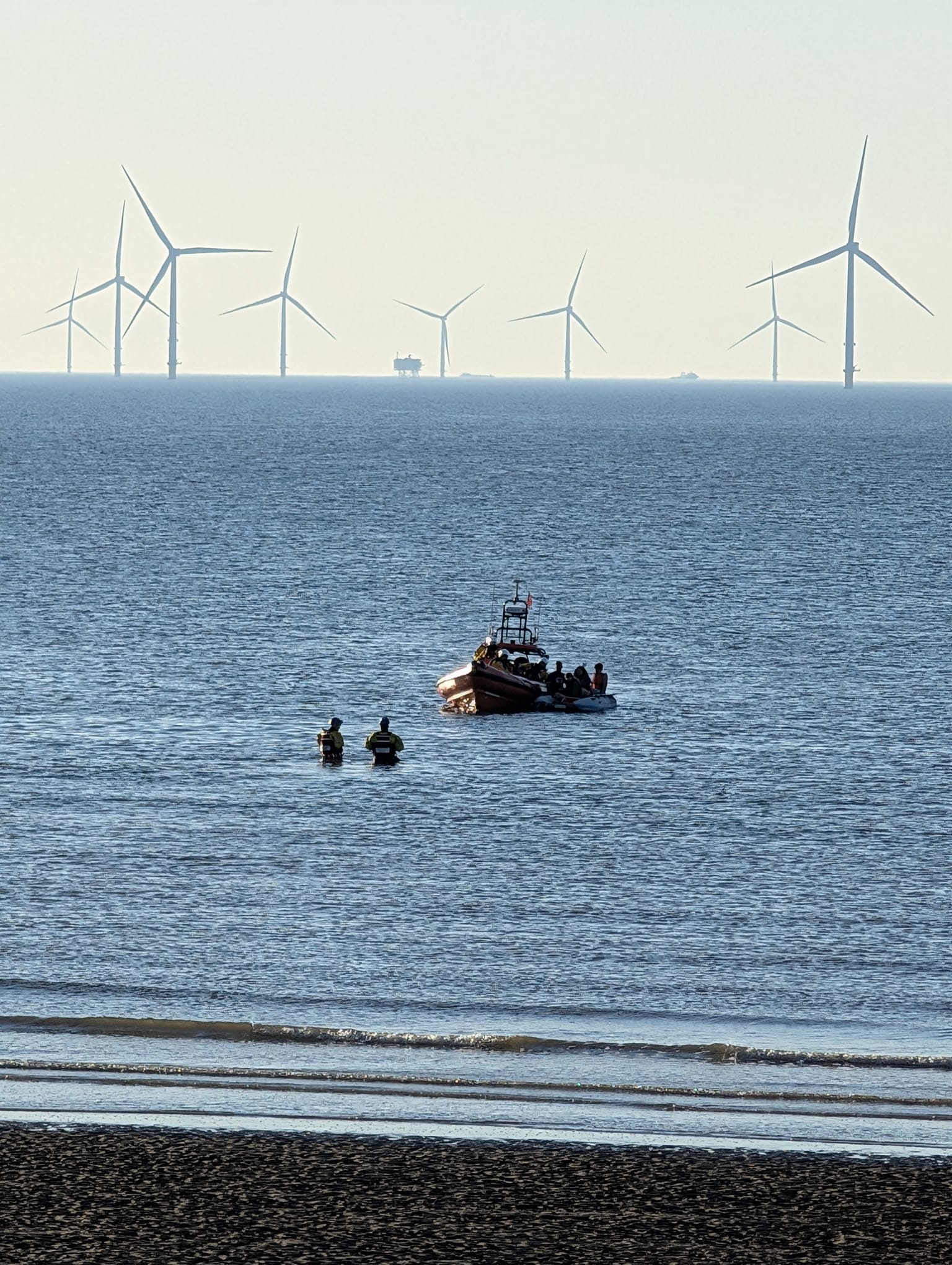 A portrait shot of the Mersey, with wind farms visible in the background, and beach in the foreground. Two Coastguard rescue units head out, wading in the water, to greet New Brighton RNLI's Atlantic-85, with the casualty vessel.

