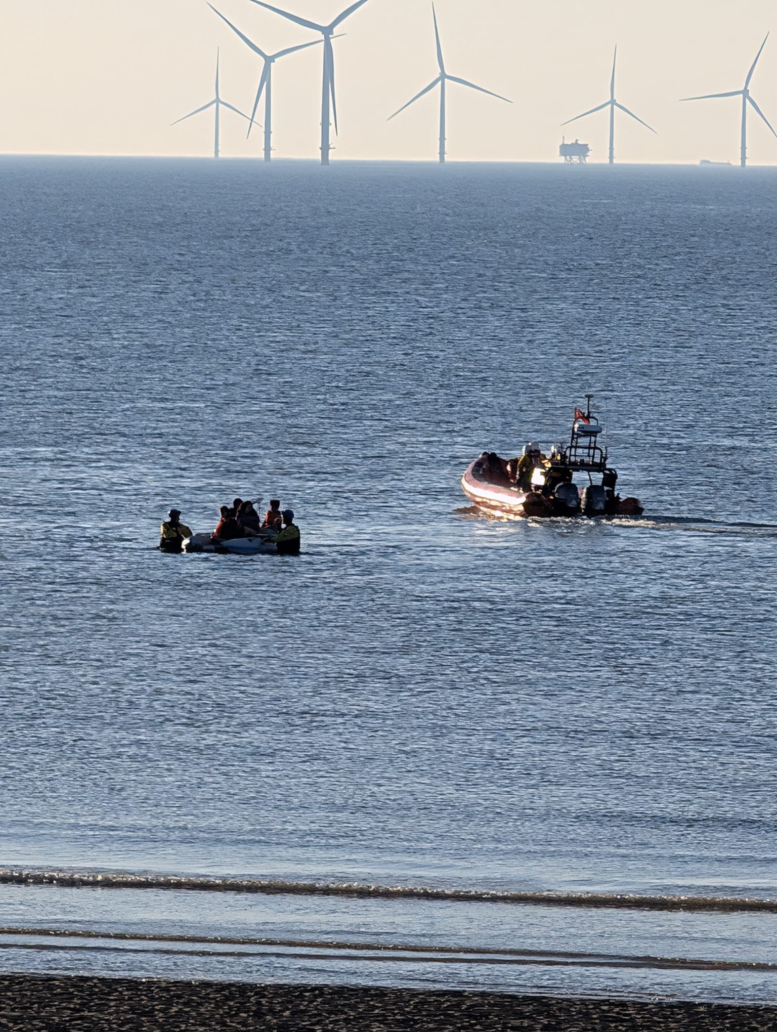 Wind farm in the background, small strip of beach in the foreground. The majority of the frame is dominated by water, in a portrait orientation, as New Brighton RNLI head away from the scene. Two Coastguard units can be seen assisting the small dinghy of casualties back to shore. 