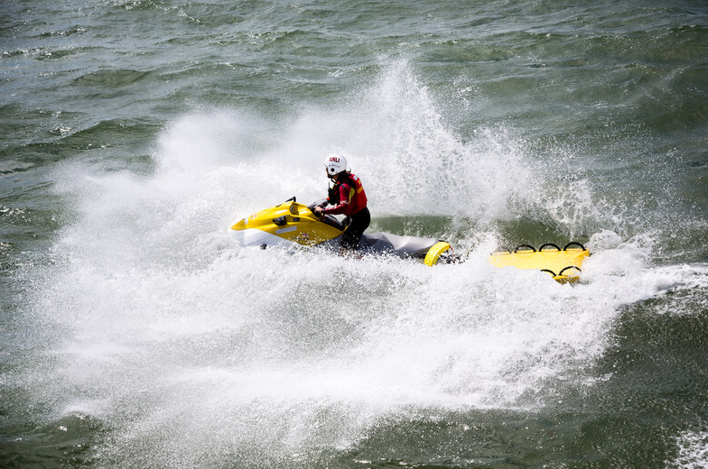 Lifeguard wearing a white helmet on a yellow and white rescue water craft crashes through the waves with a yellow rescue board attached.