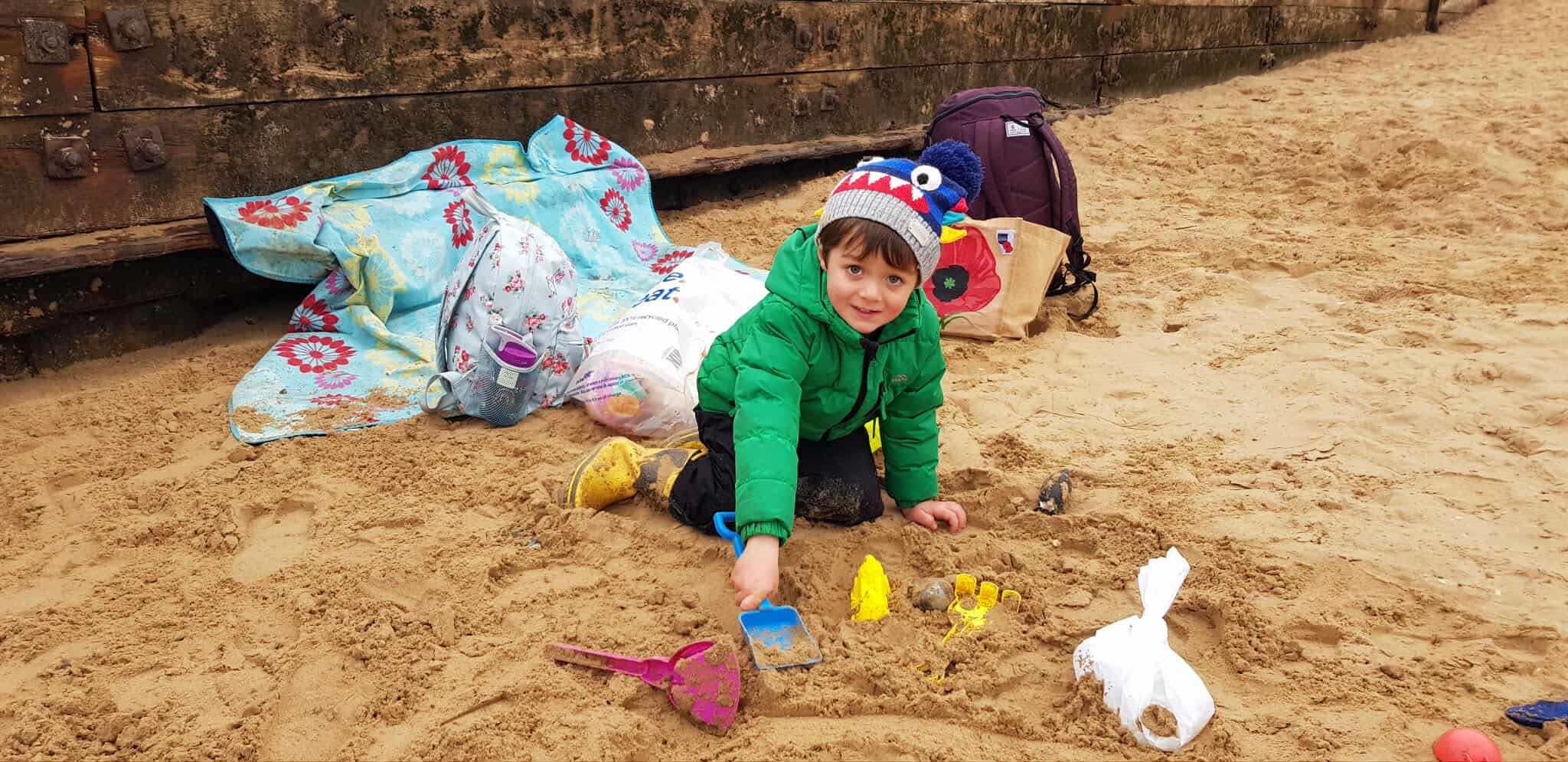 Sam in Green coat playing in the sand on Wells beach with a blue hat with moster teeth and eyes