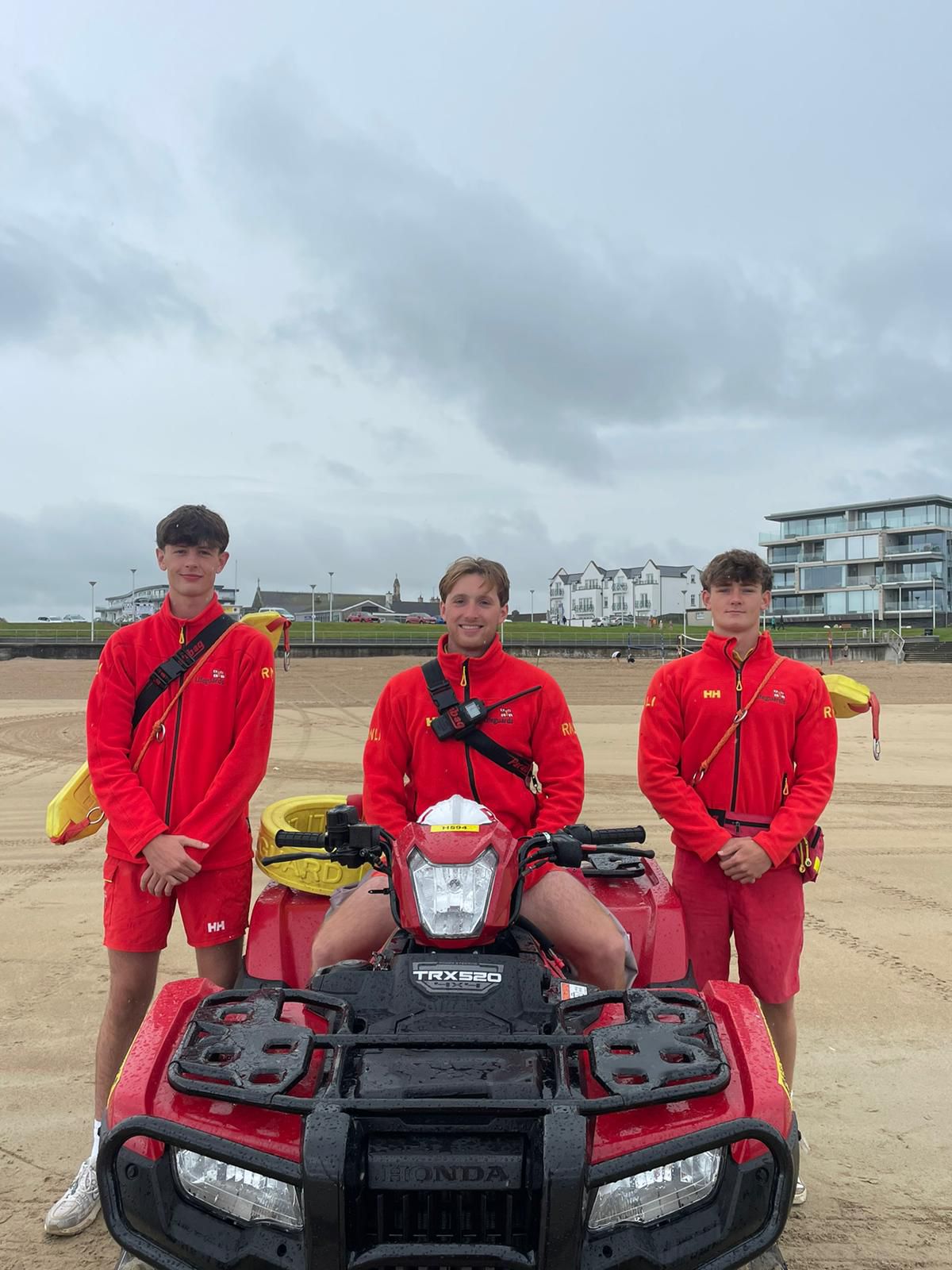 RNLI Lifeguards Oliver Knox, Cameron Leighton and Paul Richmond. Cameron is in the middle sitting on the Quad Bike with Oliver and Paul on either side. They're all in their RNLI Lifeguard gear and smiling to the camera.
