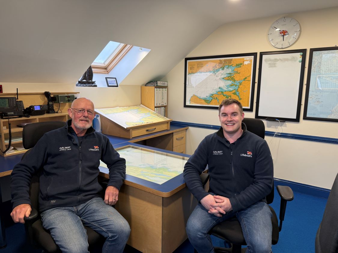 Two crew wearing navy RNLI jackets sitting in the ops room of Galway Lifeboat Station with charts and comms equipment in the background
