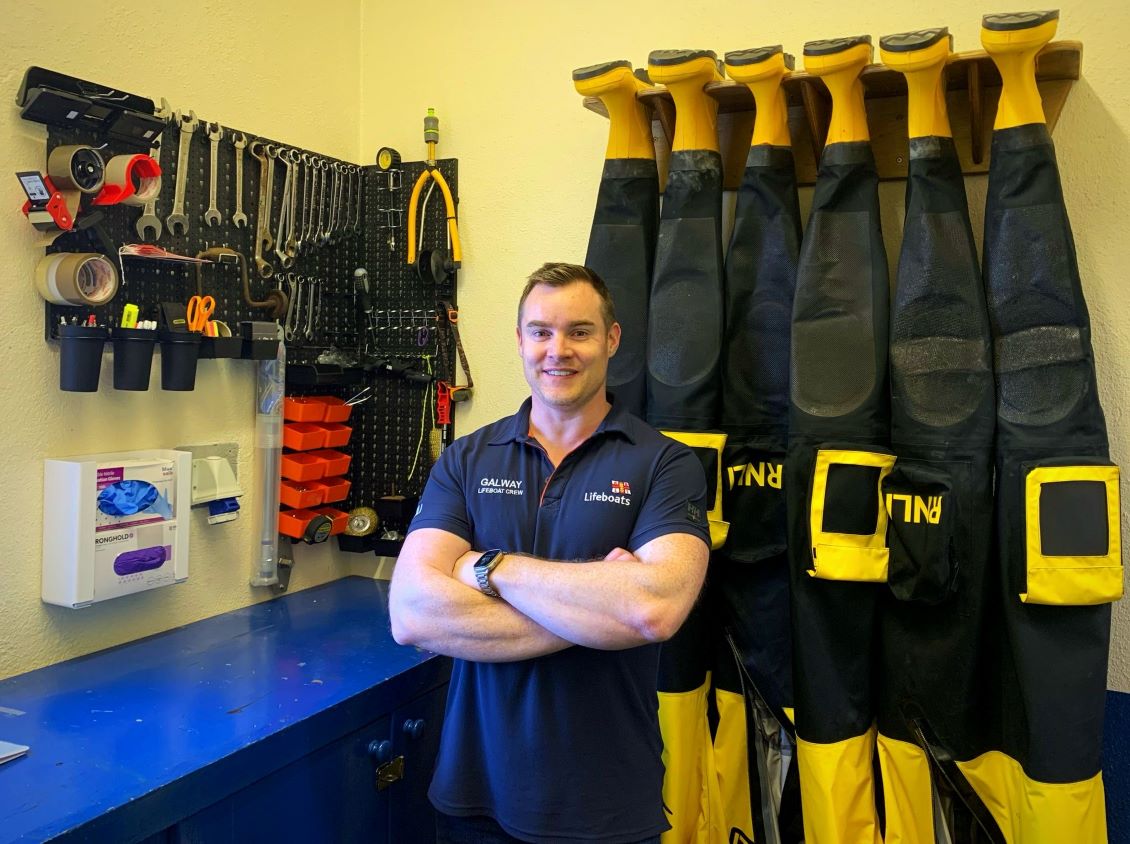 Male volunteer with arms folded facing the camera in the mechanic's workshop with three drysuits hanging upside down and a panel of tools on the walls in the background