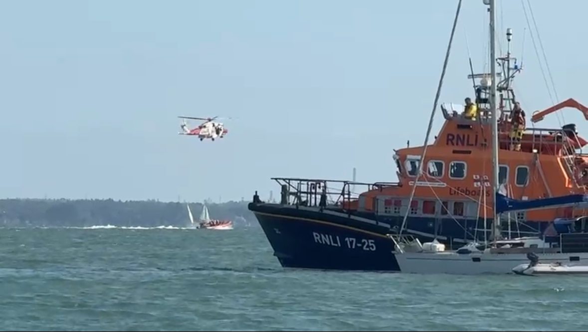 Yarmouth RNLI's ALB is to to the right of the frame with a yacht beside it in the foreground. A helicopter is seen overhead in the background.