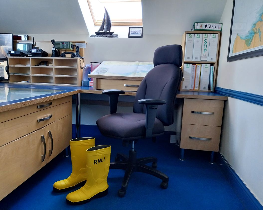 Inside the ops room with an empty office chair in front of two desks with files and comms equipment in the background. On the ground in front of the chair are two empty RNLI yellow wellies