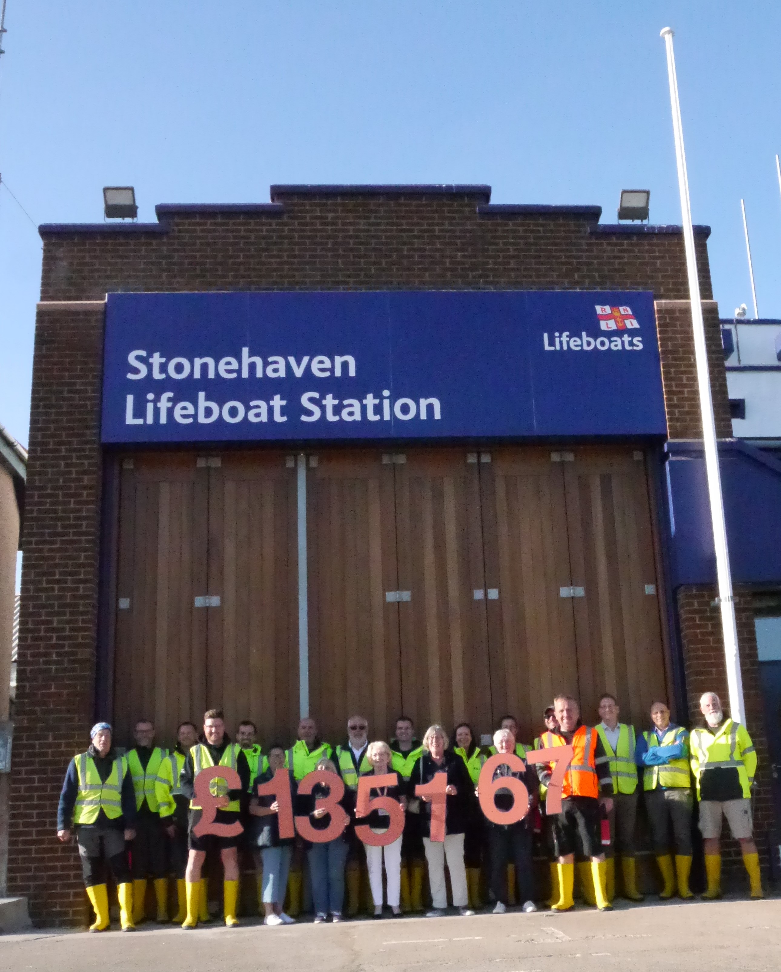 Stonehaven RNLI volunteer crew standing in front of the new station boat hall large wood doors holding the total raised by the appeal